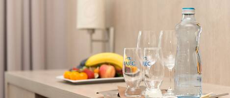 Habitación de hotel con mostrador de madera, dos vasos, una botella de agua y un plato de frutas con plátanos y manzanas.