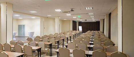 A conference room with rows of tables and chairs, a projector screen, and a stage at the front.