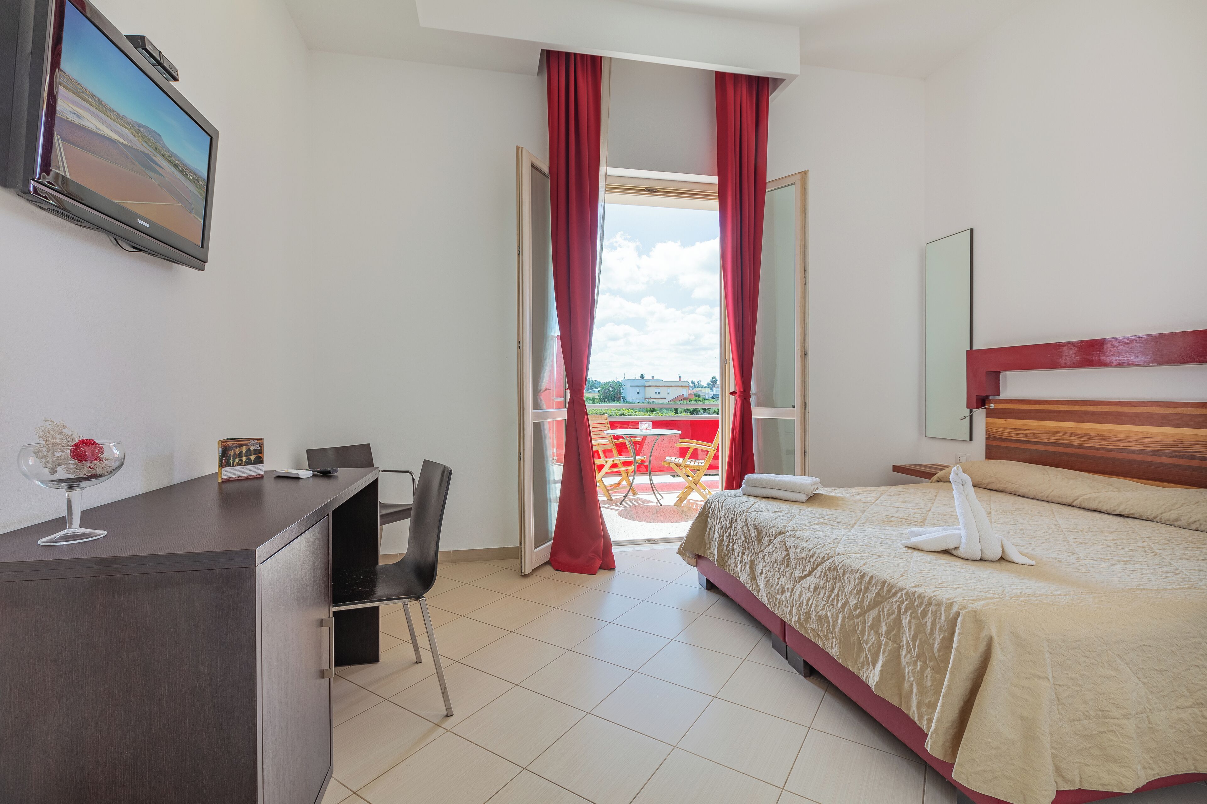 Una camera d'albergo con un letto, una scrivania, una sedia e un balcone con tende rosse.