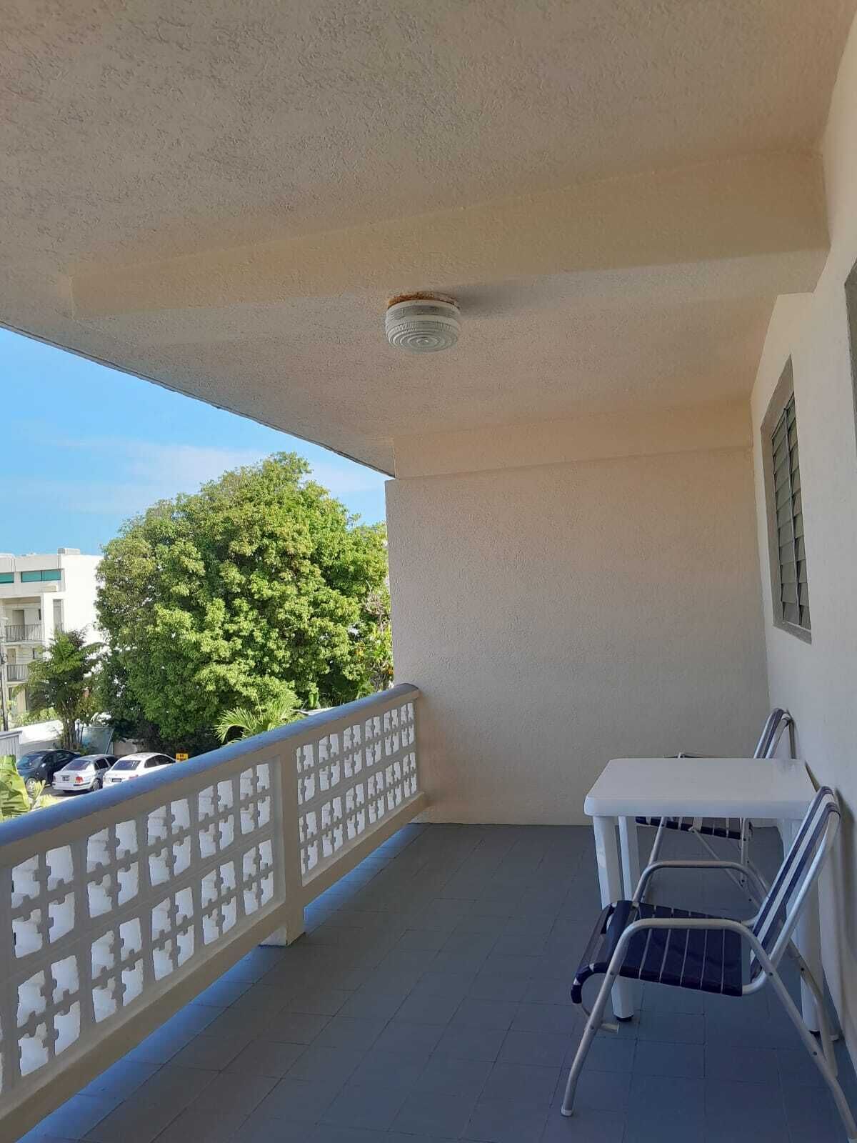 Un balcón con una mesa y una silla blancos, barandilla blanca y vistas a árboles y edificios.