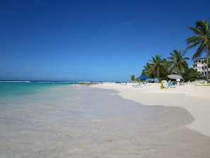 A beach with clear turquoise water, white sand, and palm trees. There are lounge chairs and umbrellas along the shore.