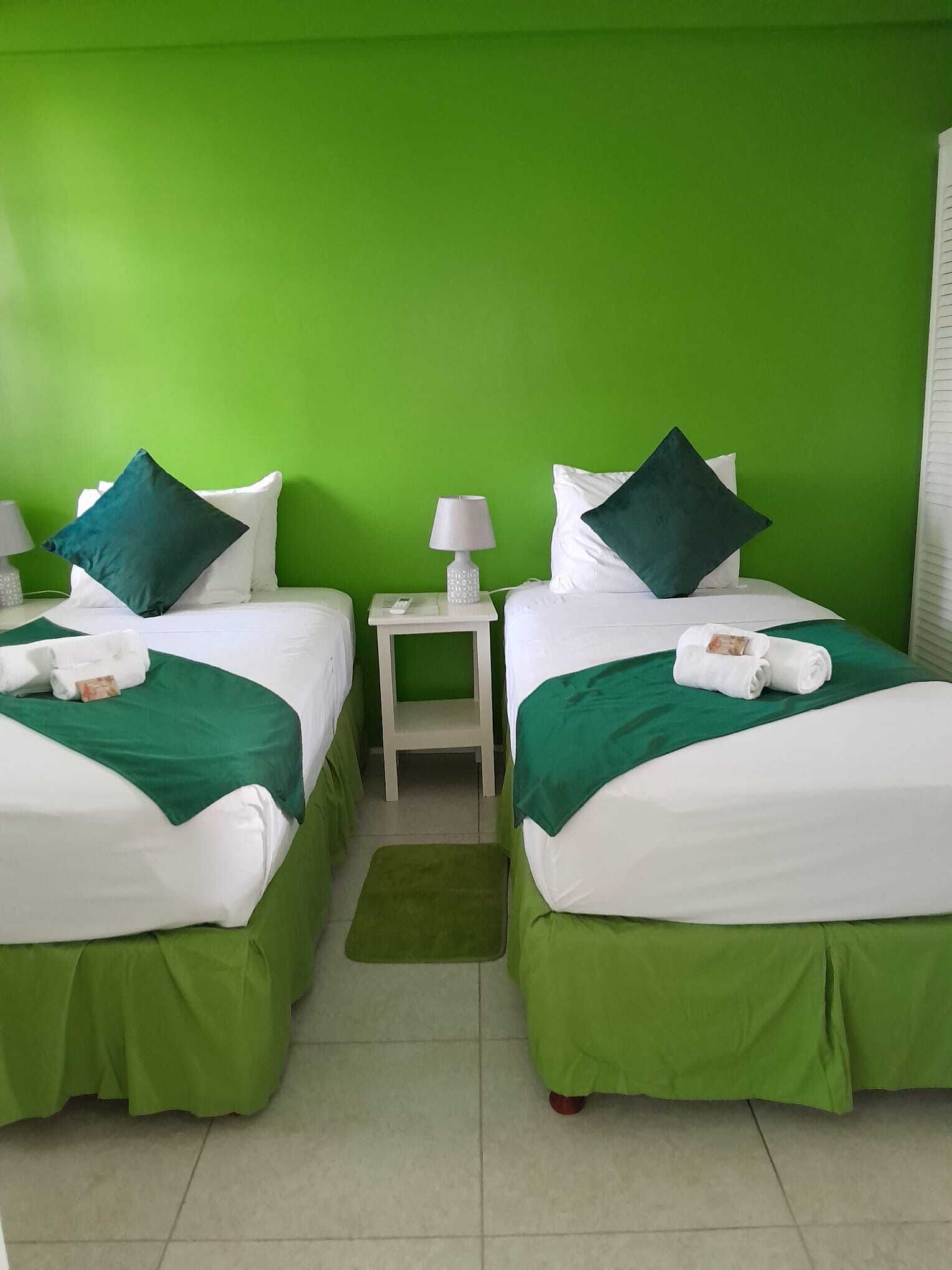A hotel room with two beds, each with green bedding and white pillows, a small bedside table with a lamp, and a green rug on the floor.