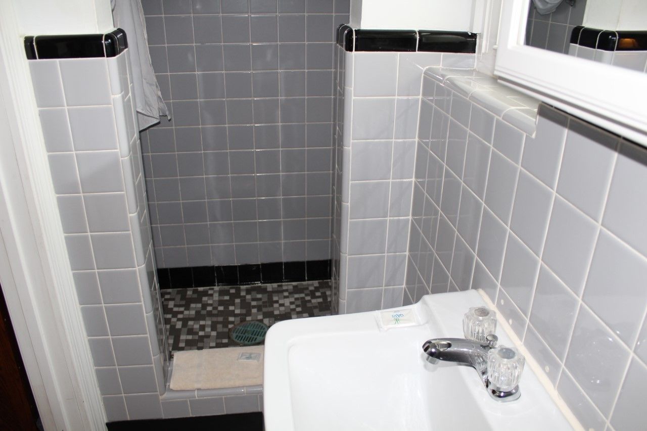 Un baño con paredes de azulejos grises, un lavamanos blanco con dos grifos y una zona de ducha con suelo de azulejos.