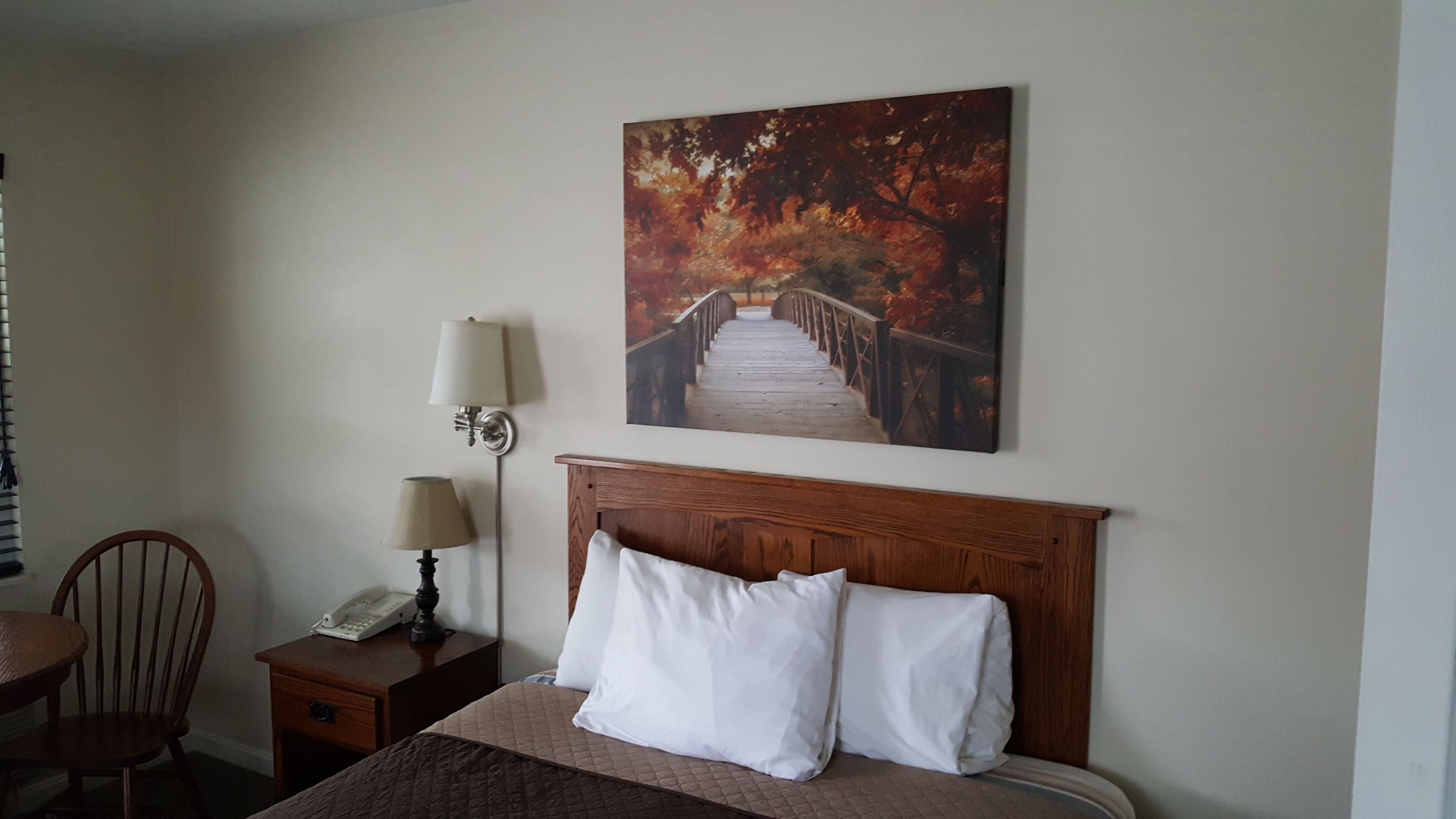 A hotel room with a wooden headboard, a bed with white pillows, a nightstand with a lamp, a chair, and a painting of a bridge.