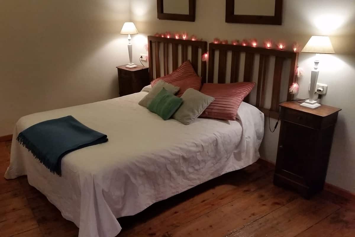A neatly made bed with white linens, a blue throw blanket, and several pillows in various colors. There are two bedside tables with lamps, and the room has wooden flooring.