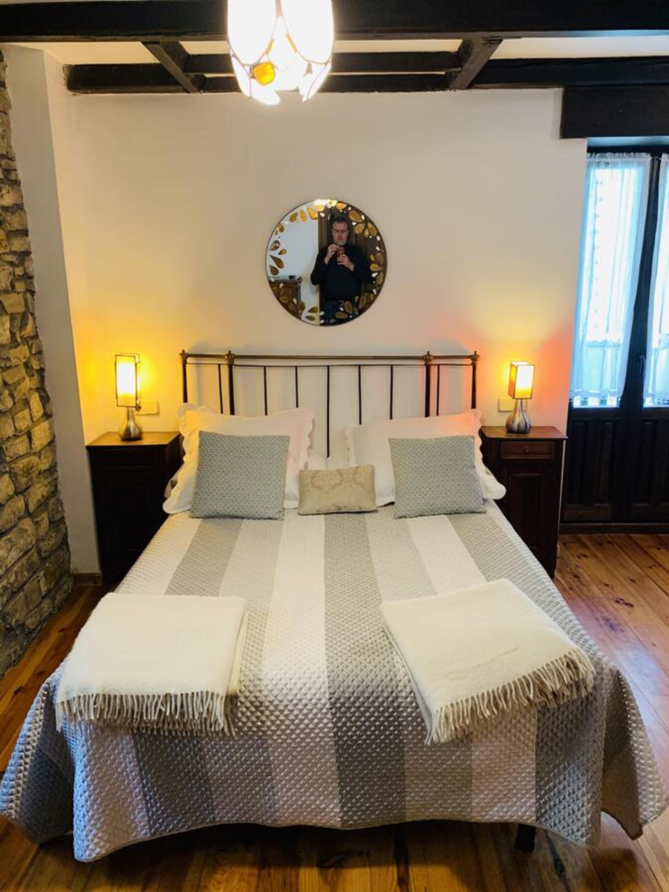 Quarto com uma cama, duas mesas de cabeceira, um espelho redondo e uma parede de pedra.