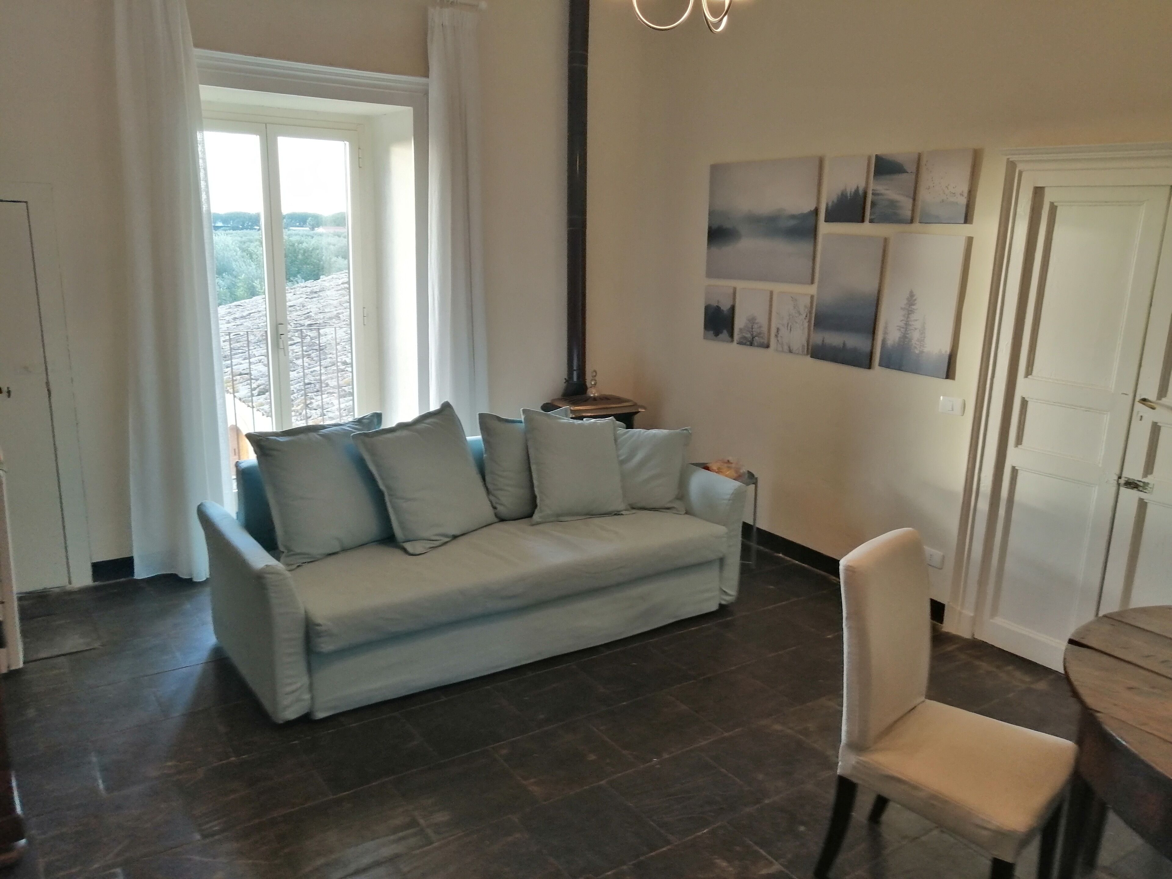 A living room with a light blue sofa, a wooden coffee table, and a white chair. There is a window with white curtains, a door, and wall-mounted artwork.