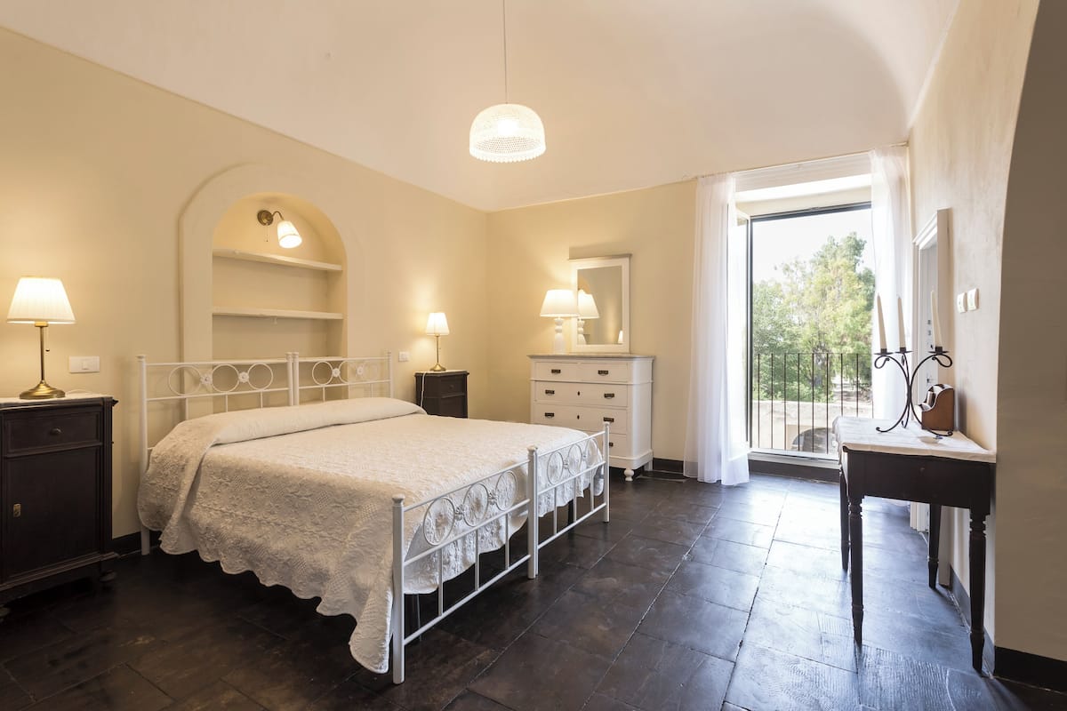 A bedroom with a bed, bedside tables, a dresser, a mirror, and a balcony with a railing.