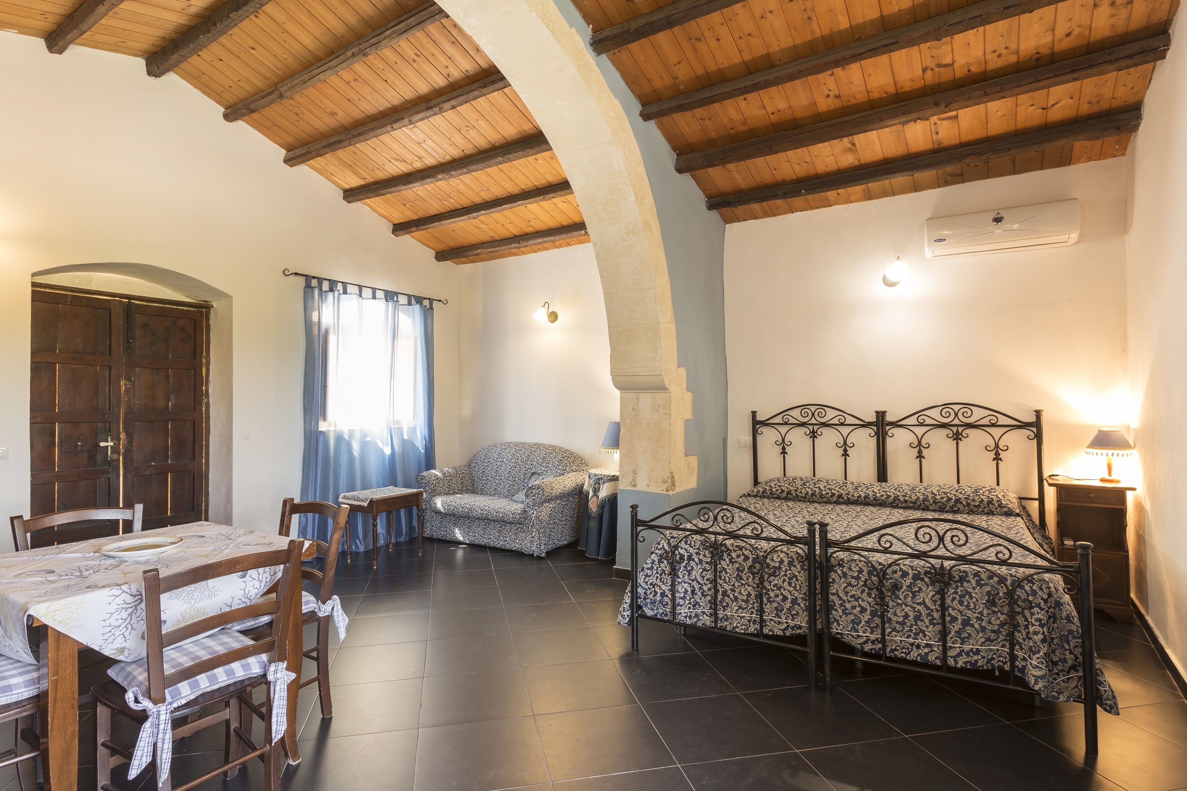 Una stanza con un letto, un tavolo da pranzo, sedie, una lampada e una finestra con tende.