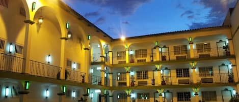 Un edificio de varios pisos con un patio, con balcones que tienen barandillas y plantas en macetas.