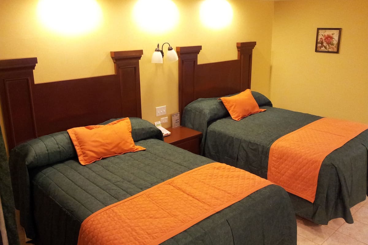 A hotel room with two beds, each with an orange quilted bedspread and matching pillows. There are wall-mounted lights above the beds and a small table beside them.