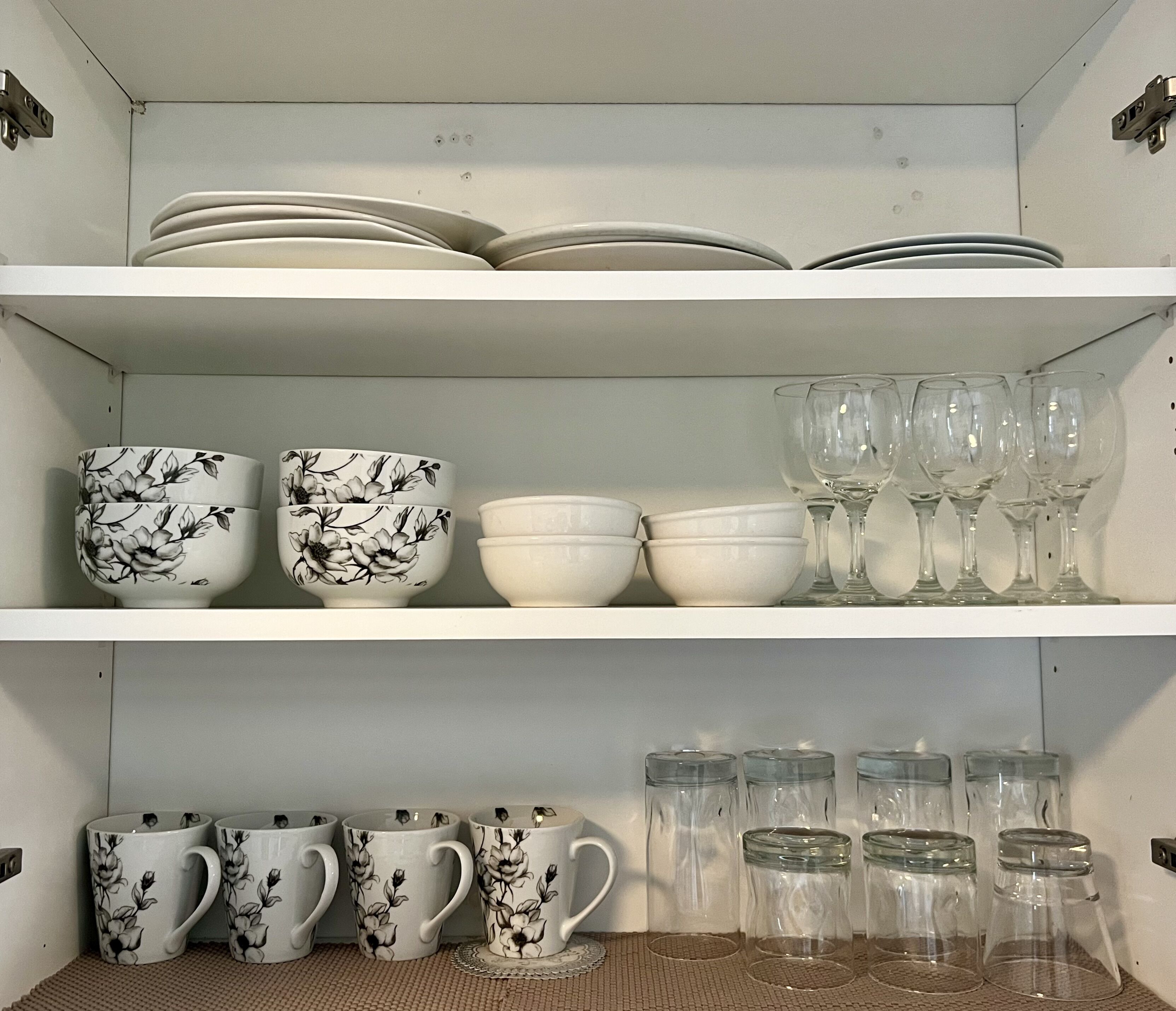 A kitchen shelf with white plates, bowls, and mugs, some with floral designs, and clear glassware.