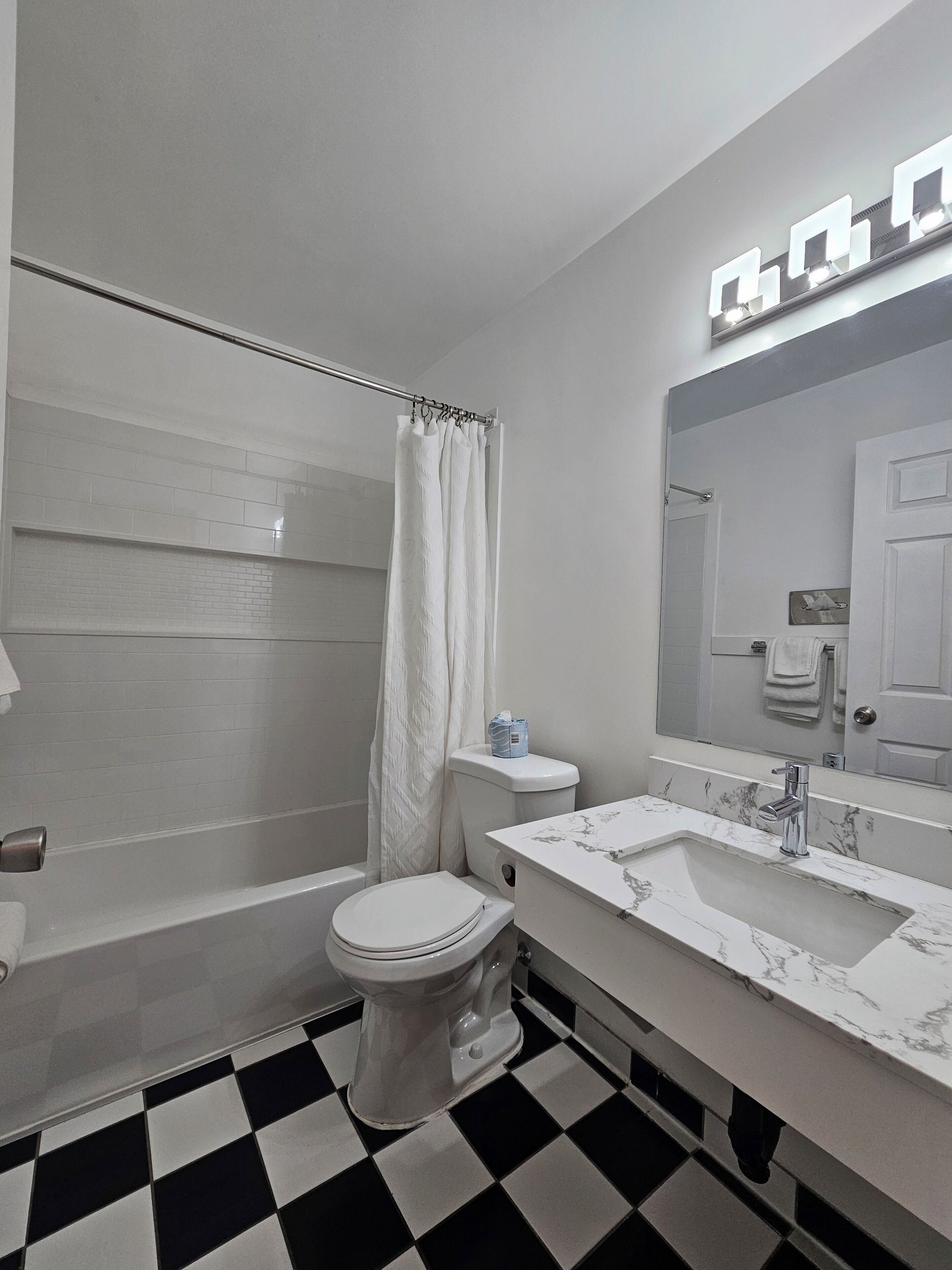 A bathroom with a white toilet, a bathtub, a shower curtain, a sink with a marble countertop, and a mirror with lights above it.