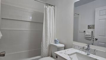 A bathroom with a white toilet, a bathtub, a shower curtain, a sink with a marble countertop, and a mirror with lights above it.