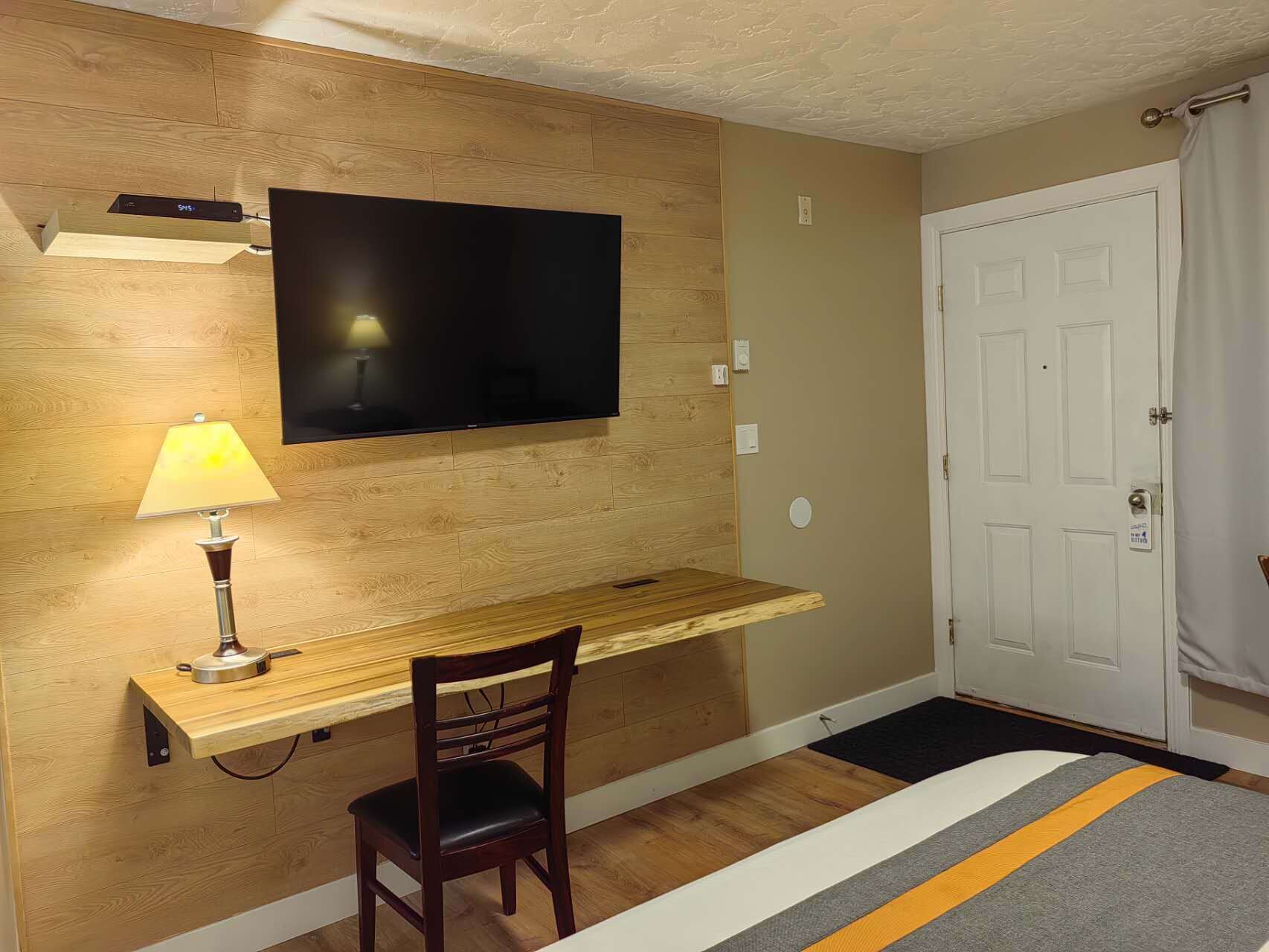 A hotel room with a wooden desk, a chair, a TV mounted on the wall, a lamp, and a door with a doormat.