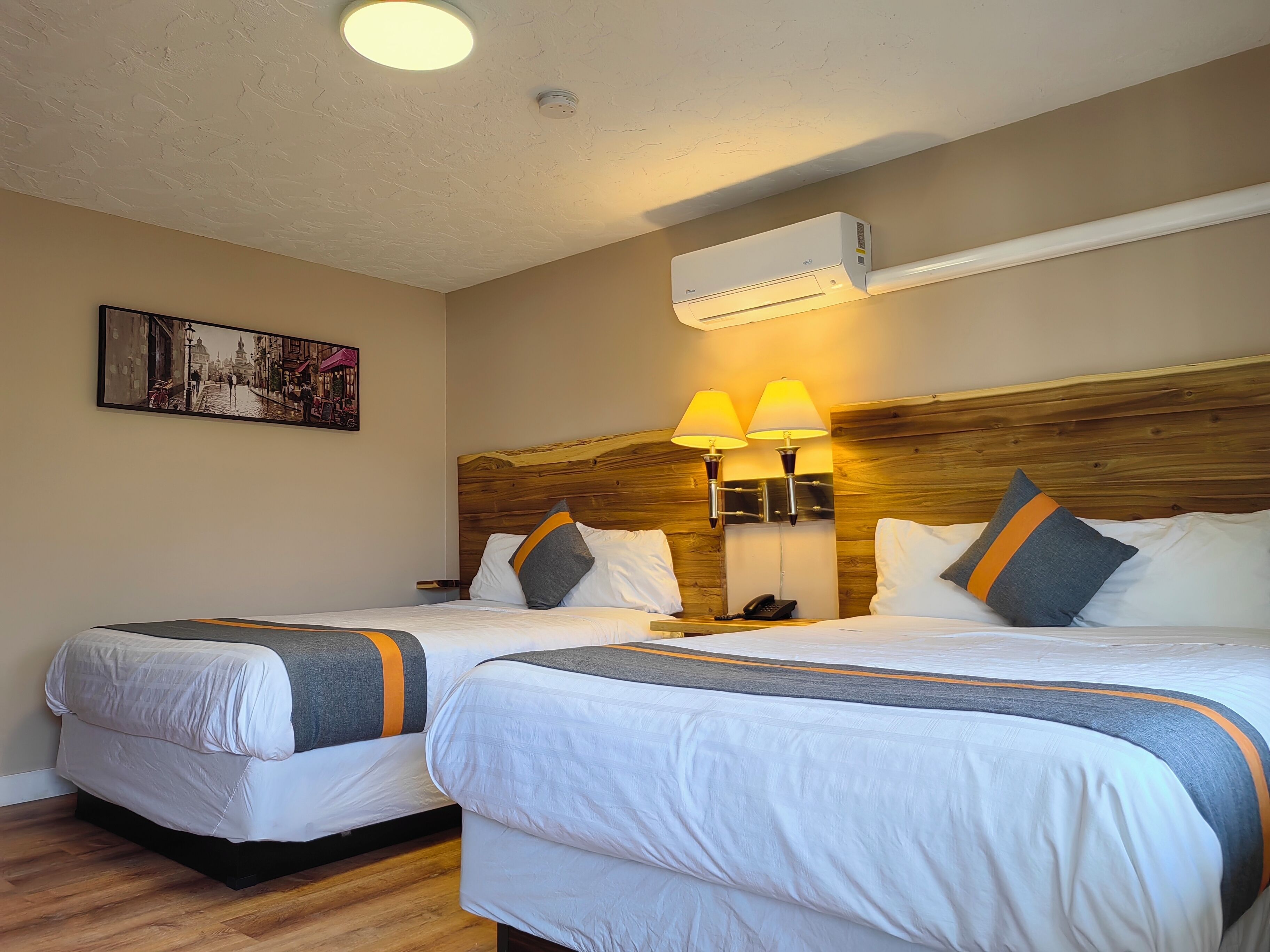 A hotel room with two beds, a wooden headboard, bedside lamps, an air conditioner, and a framed picture on the wall.