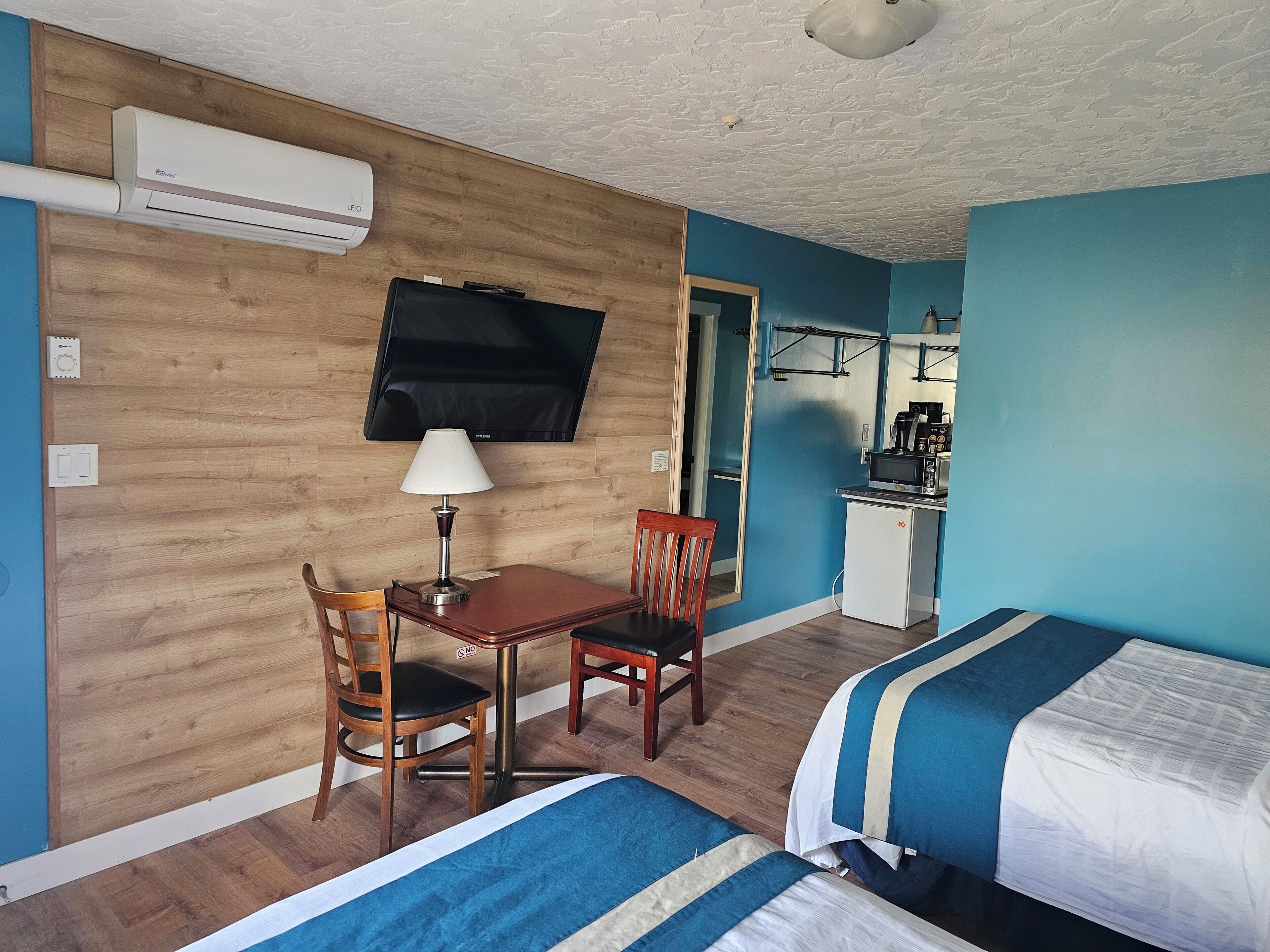 A hotel room with a wooden wall, a TV, a desk with a lamp, a chair, a bed with striped bedding, and a kitchenette with a microwave.