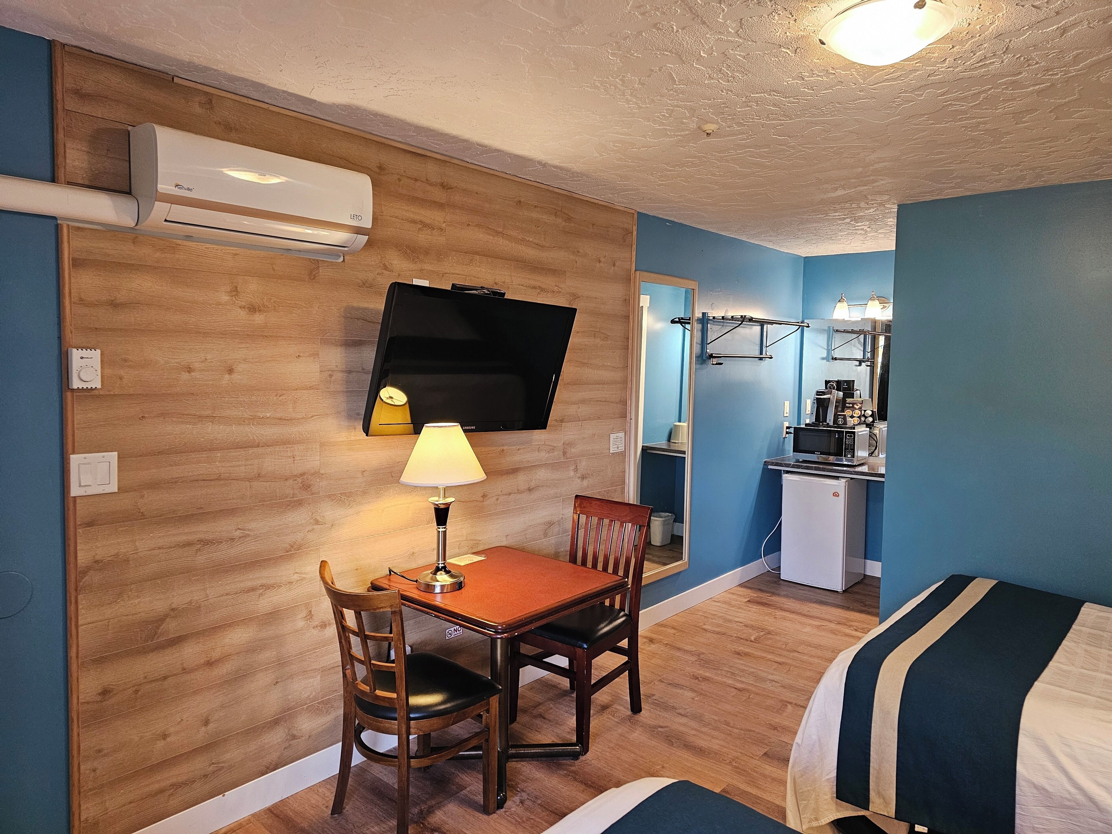 A compact hotel room with a wooden wall, a flat-screen TV, a small kitchenette, a dining table with a chair, and a bed with striped bedding.