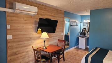 A compact hotel room with a wooden wall, a flat-screen TV, a small kitchenette, a dining table with a chair, and a bed with striped bedding.