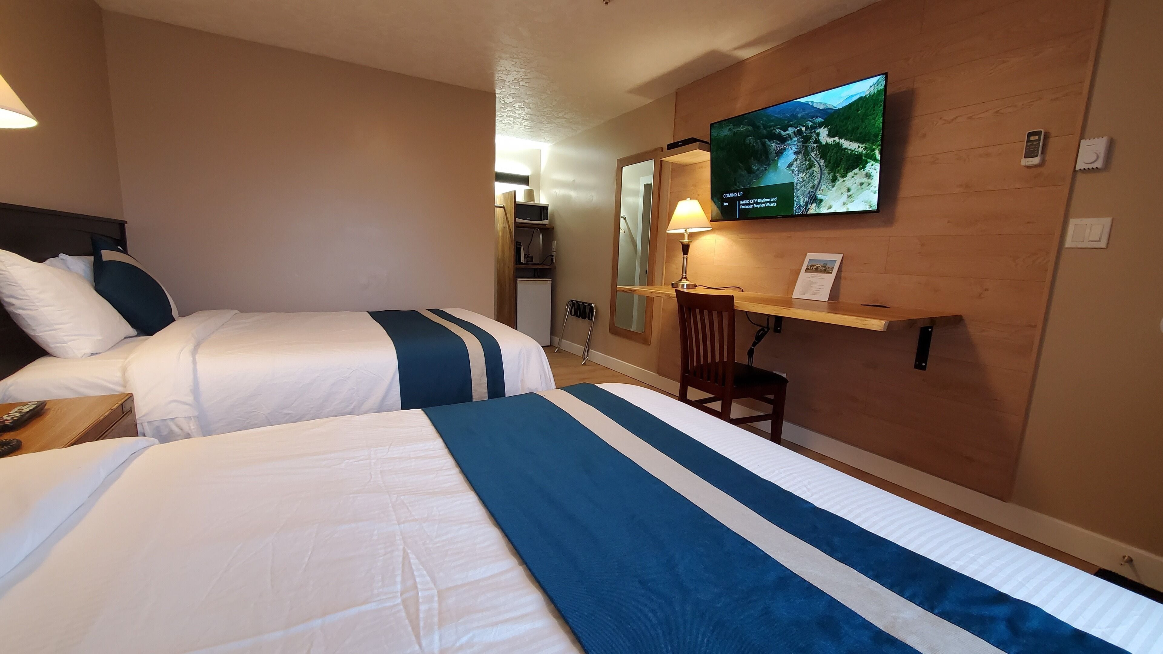 A hotel room with two beds, a desk, a TV mounted on the wall, and a small shelf with a remote control.