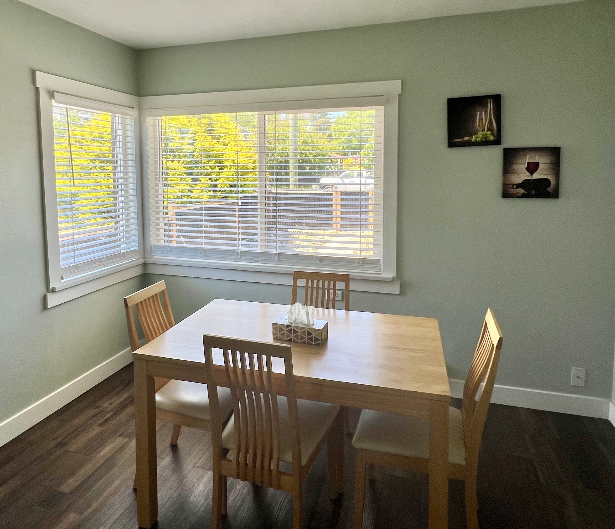 A dining room with a wooden table and chairs, large windows with blinds, and a view of a backyard.