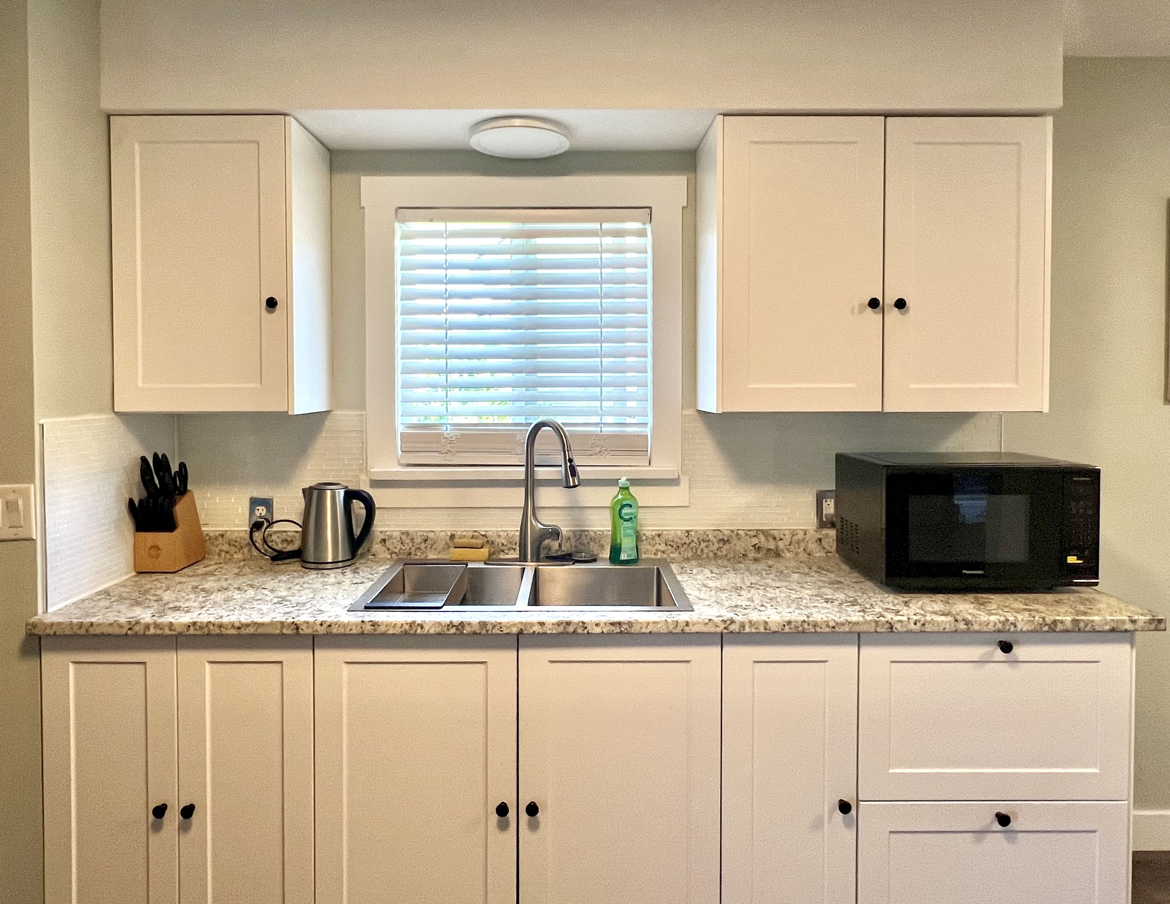 A kitchen with white cabinets, a stainless steel sink, a window with blinds, a microwave, and a knife block.