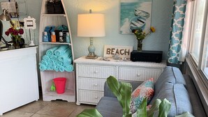 A cozy living room with a sofa, a coffee table, and a plant. There is a small white shelf with various items and a lamp. A painting of boats hangs on the wall. - The Darlington Cottages (Myrtle Beach)