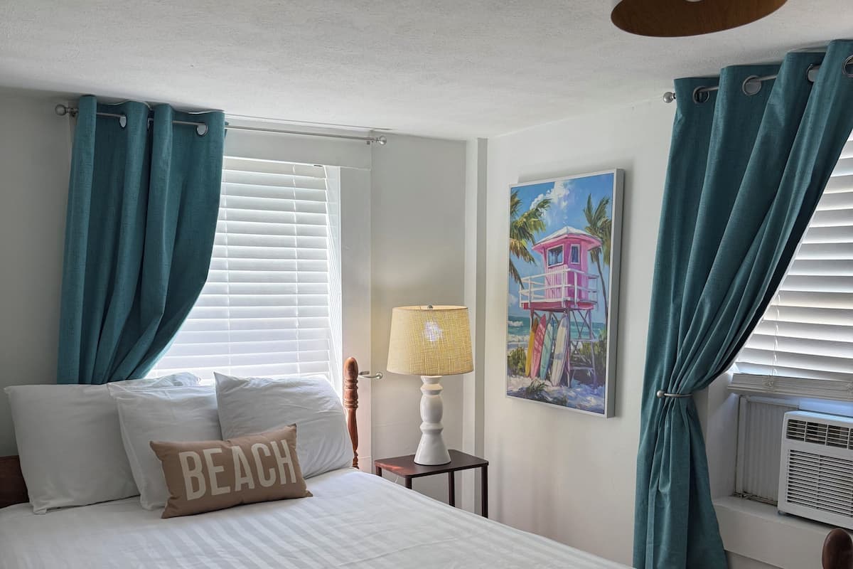 A neatly made bed with a pillow labeled 'BEACH,' a bedside lamp, a painting of a beach scene, and teal curtains.