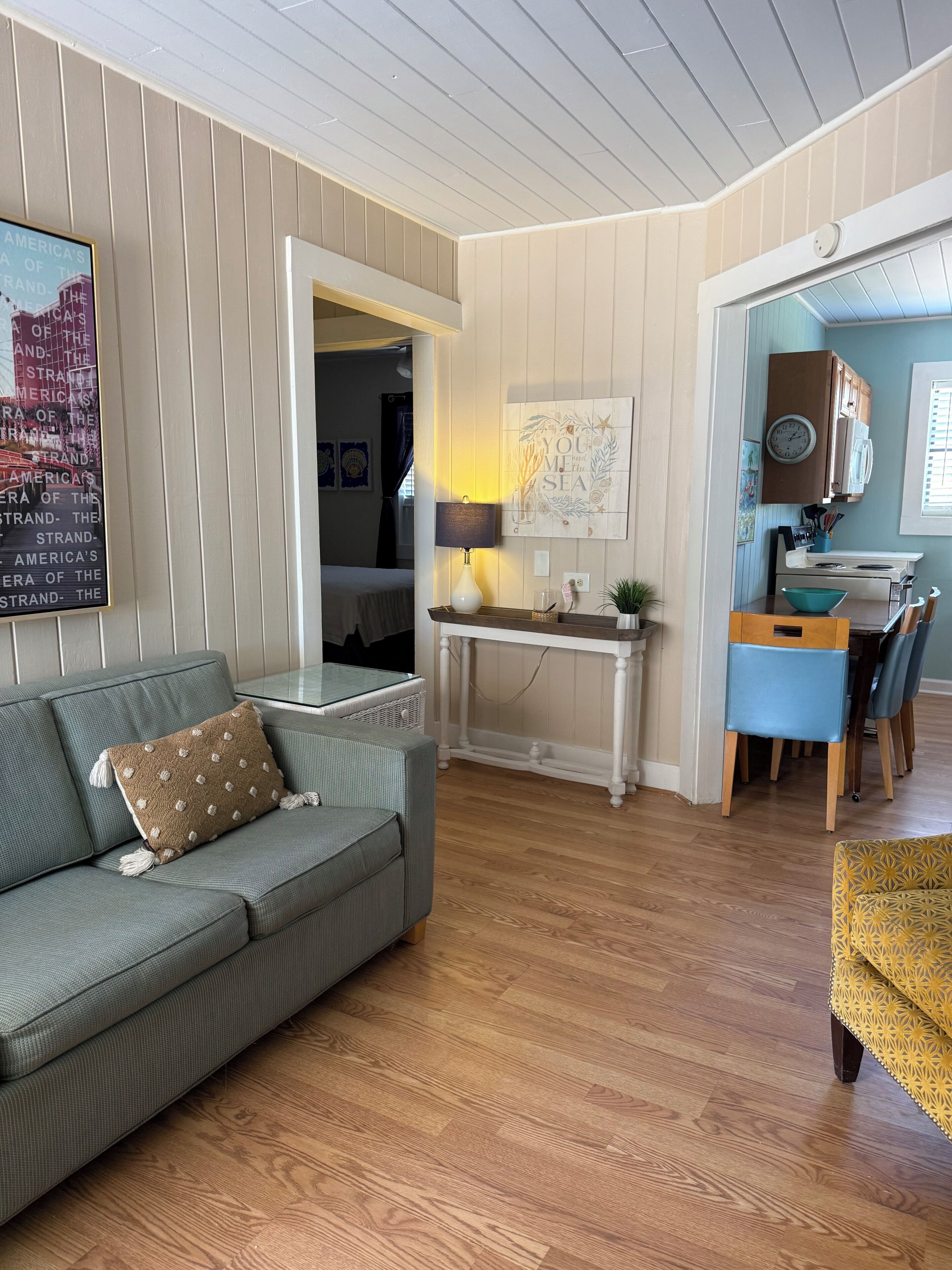 A living room with a sofa, a dining area with a table and chairs, a kitchen area with cabinets and a clock, and a hallway leading to another room.