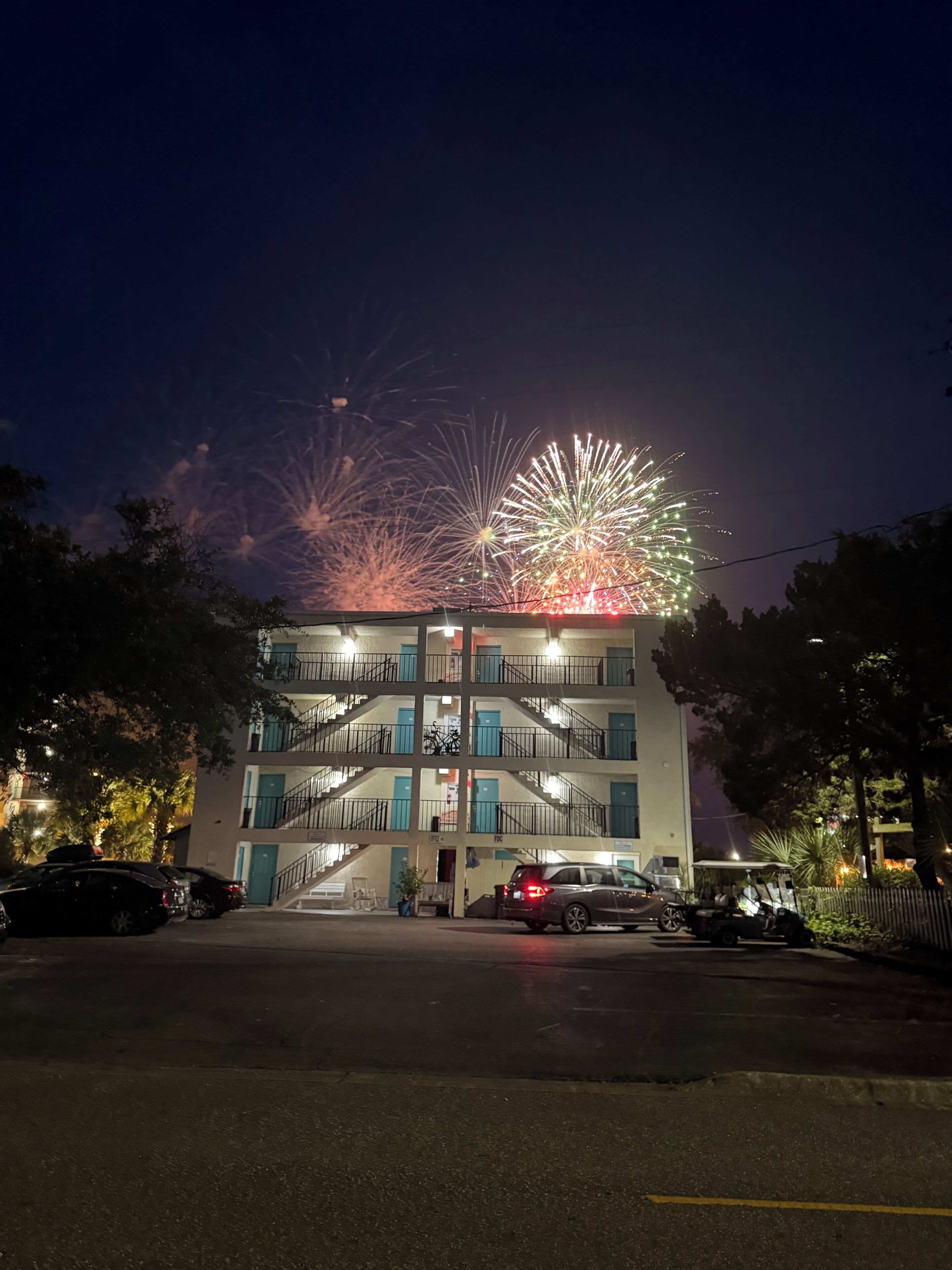 Ein mehrstöckiges Gebäude mit einer zentralen Treppe, einem Parkplatz mit Autos und einem Feuerwerk am Himmel.