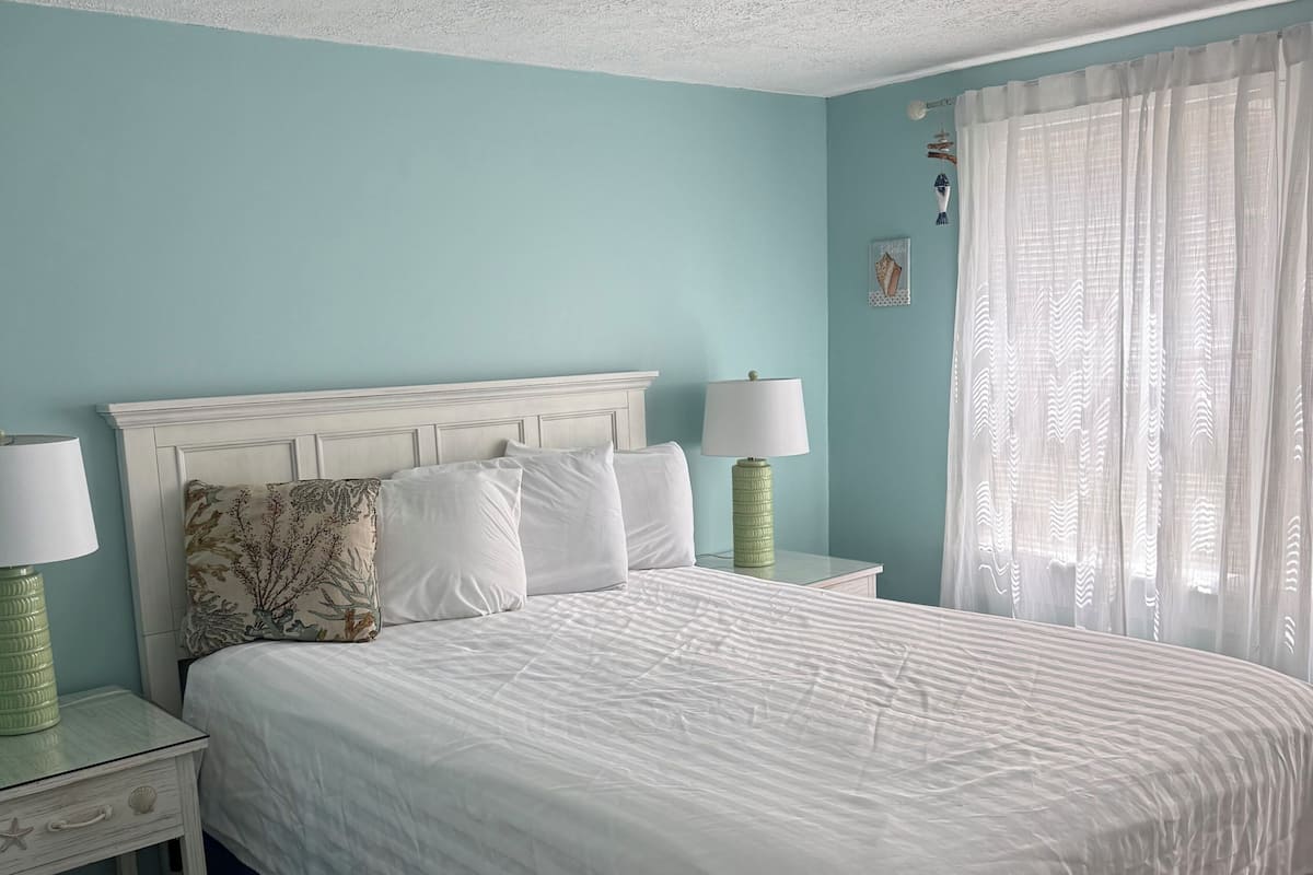 A bedroom with a bed, nightstands, a ceiling light, a window with curtains, and a wall decoration.