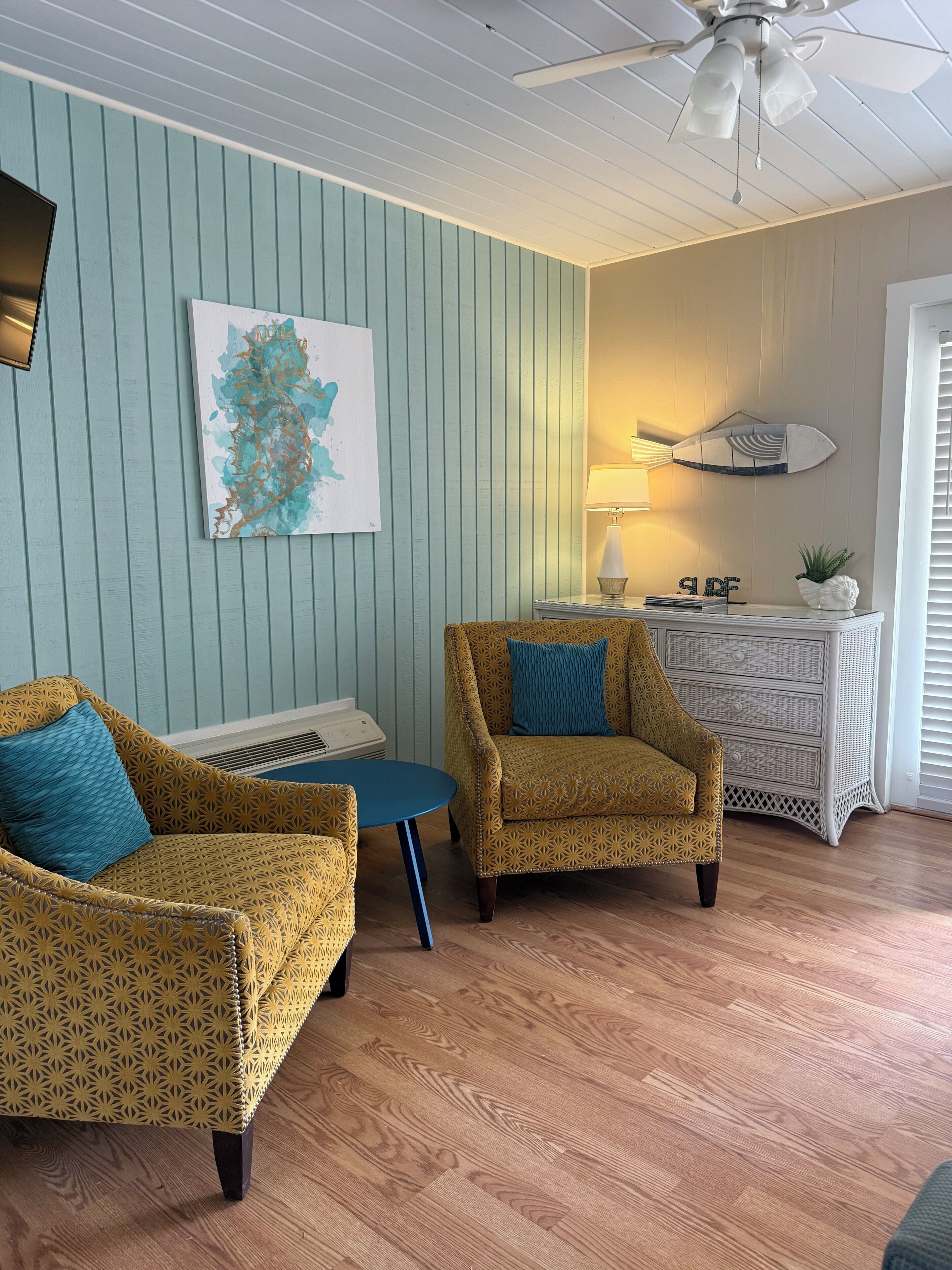 A room with two patterned armchairs, a small round table, a wall-mounted lamp, a decorative fish-shaped wall art, and a white console table with a plant.