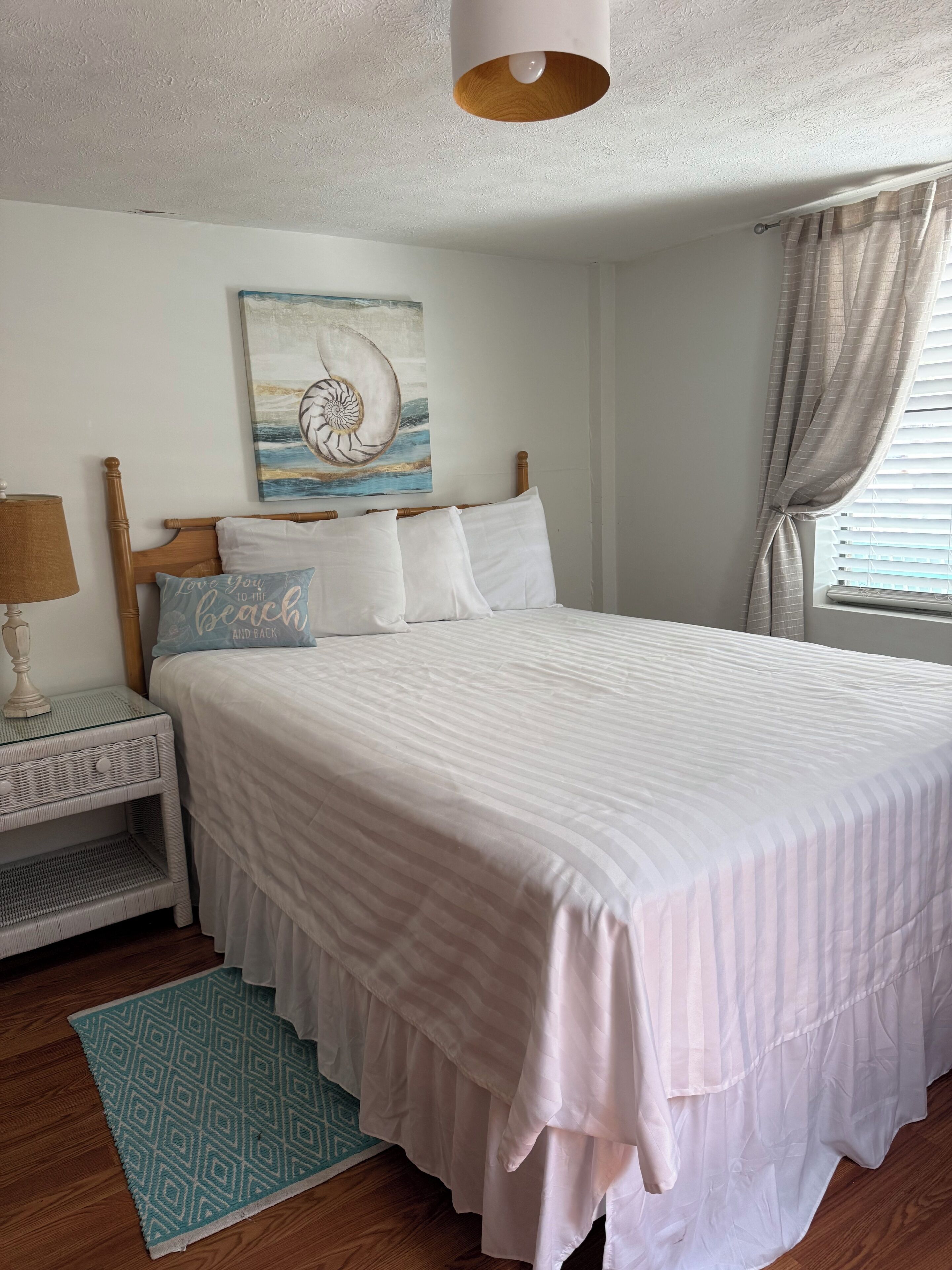 A neatly made bed with a white bedspread, a wooden headboard, a wall-mounted painting, a bedside lamp, and a window with curtains.