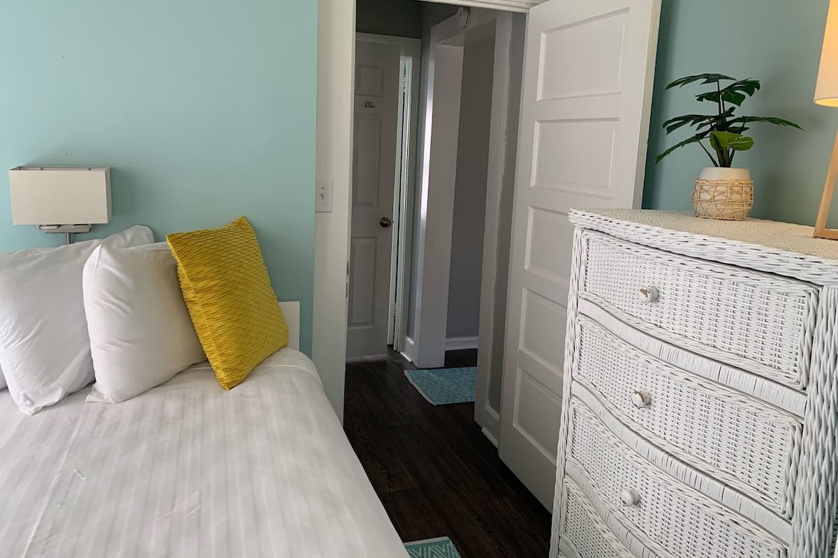 A bedroom with a bed, a white dresser, a plant, and a door leading to another room.
