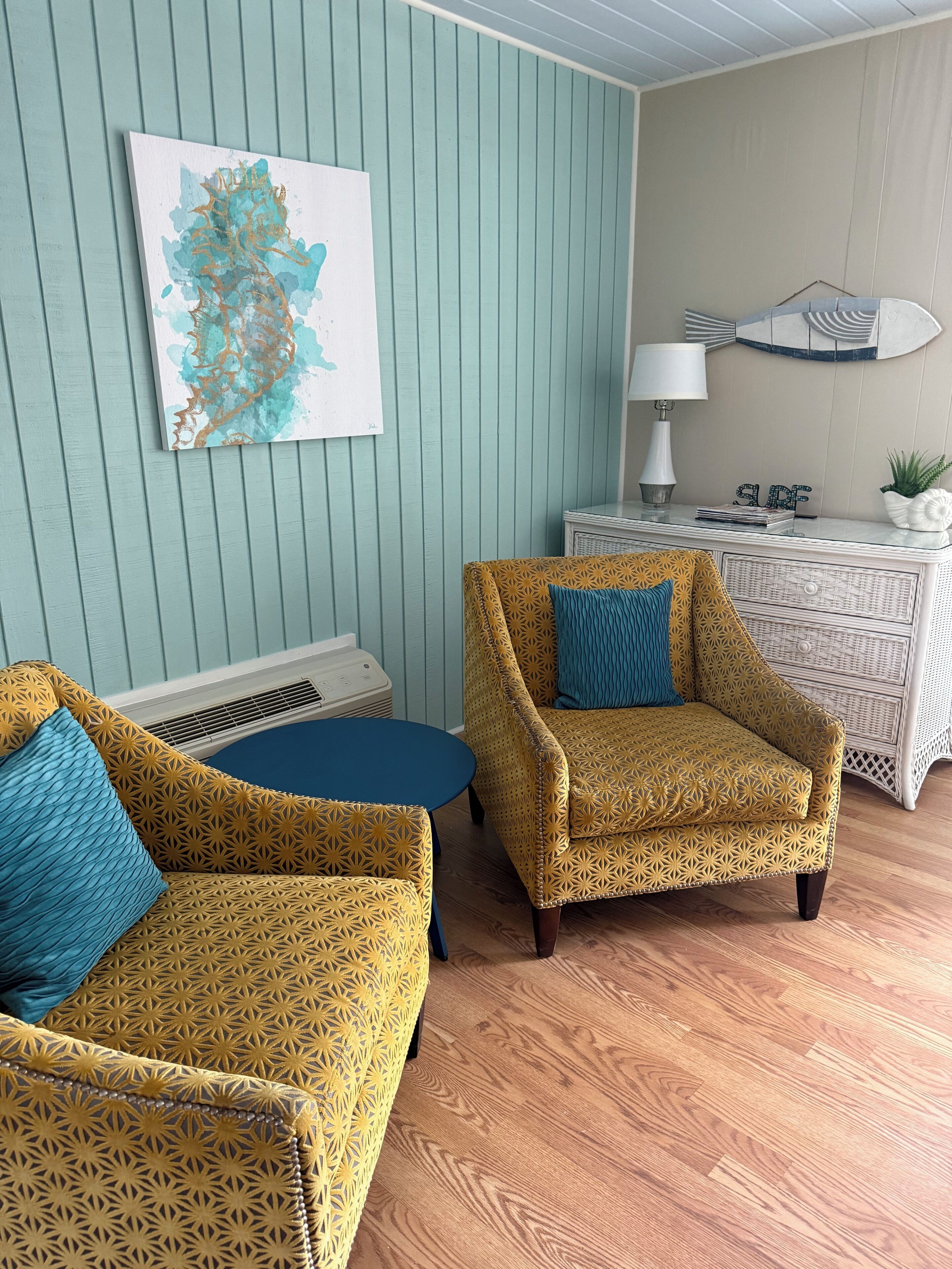 Een kamer met een sofa met een patroon, een klein rond tafeltje, een decoratieve stoel, een kunstwerk aan de muur, een witte dressoir en een lamp.