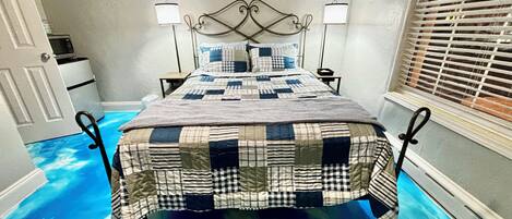 Un dormitorio con cama, dos lámparas de noche, una mesita de noche, una obra de arte en la pared y una ventana con persianas.