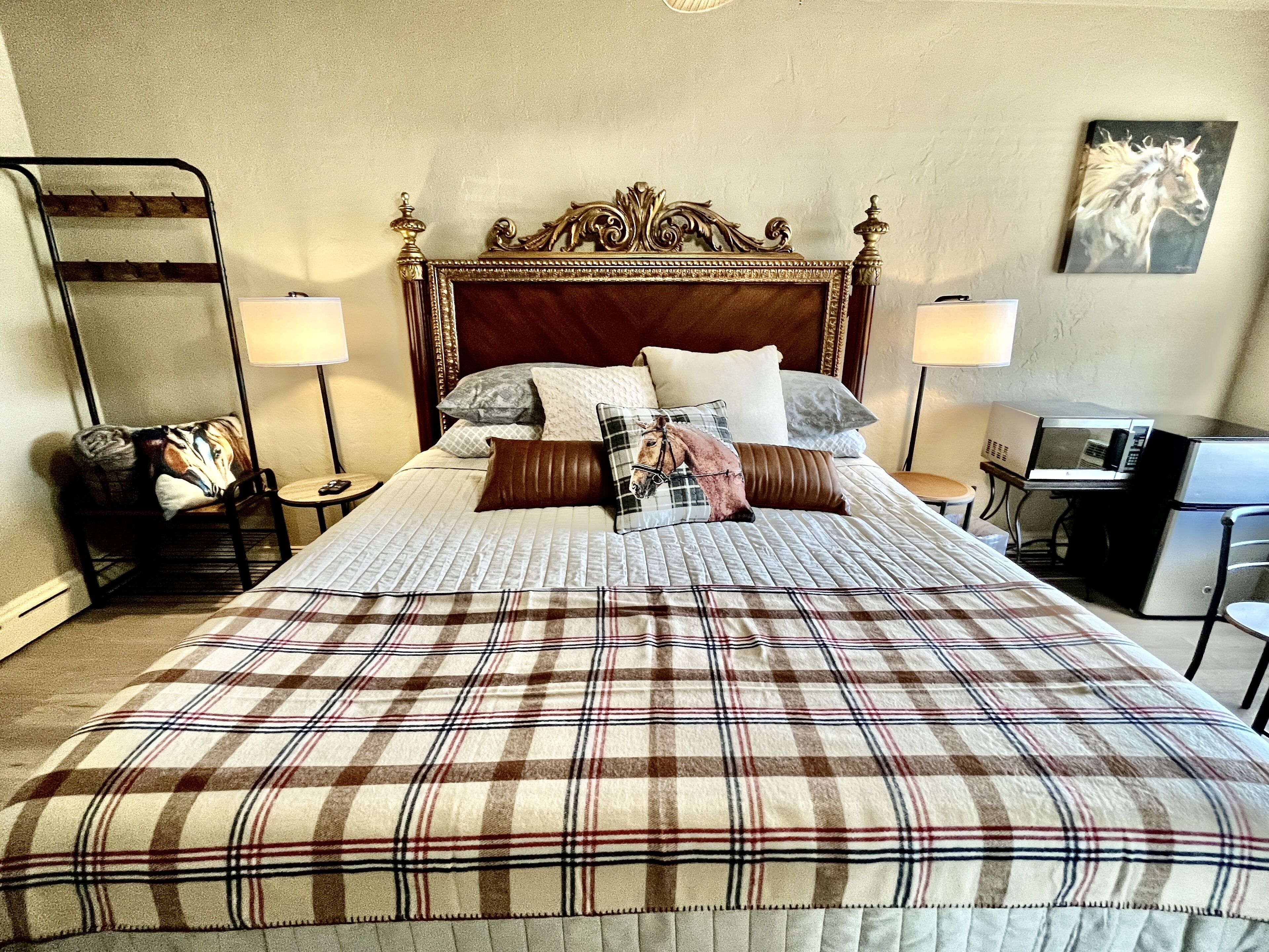 A neatly made bed with a plaid bedspread, a wooden headboard, and multiple pillows. There is a chair with a blanket, a lamp, and a framed picture on the wall.