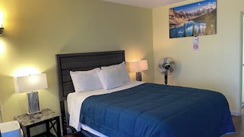 A neatly made bed with a blue comforter, a bedside lamp, a wall-mounted fan, and a painting of a mountain landscape.