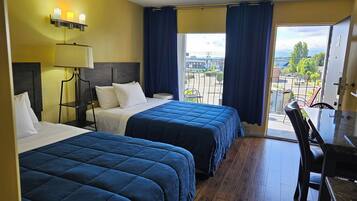 A hotel room with two beds, a desk, a chair, a lamp, and a view of the city through large windows.