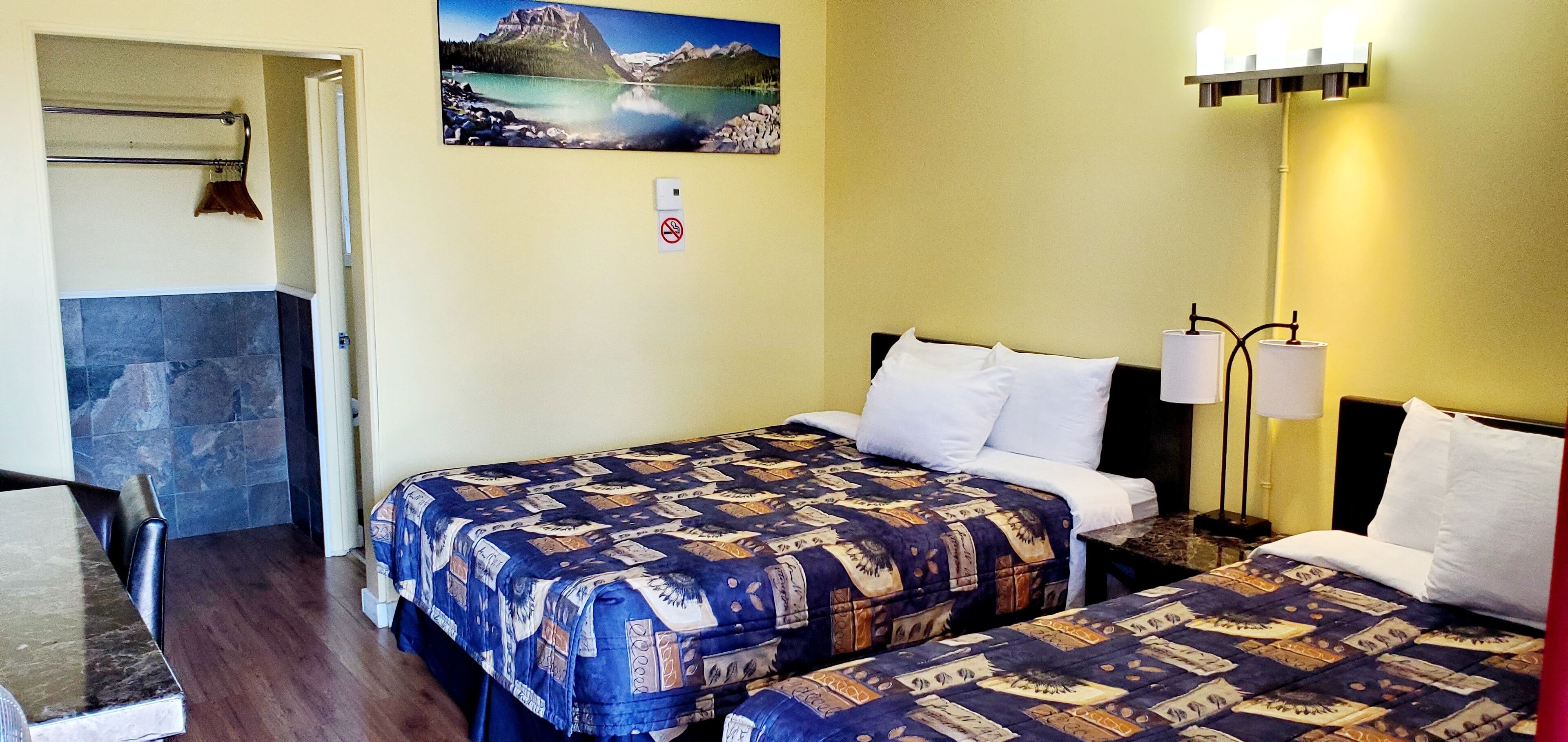 A hotel room with two beds, a painting of a mountain lake, a no-smoking sign, and a lamp.