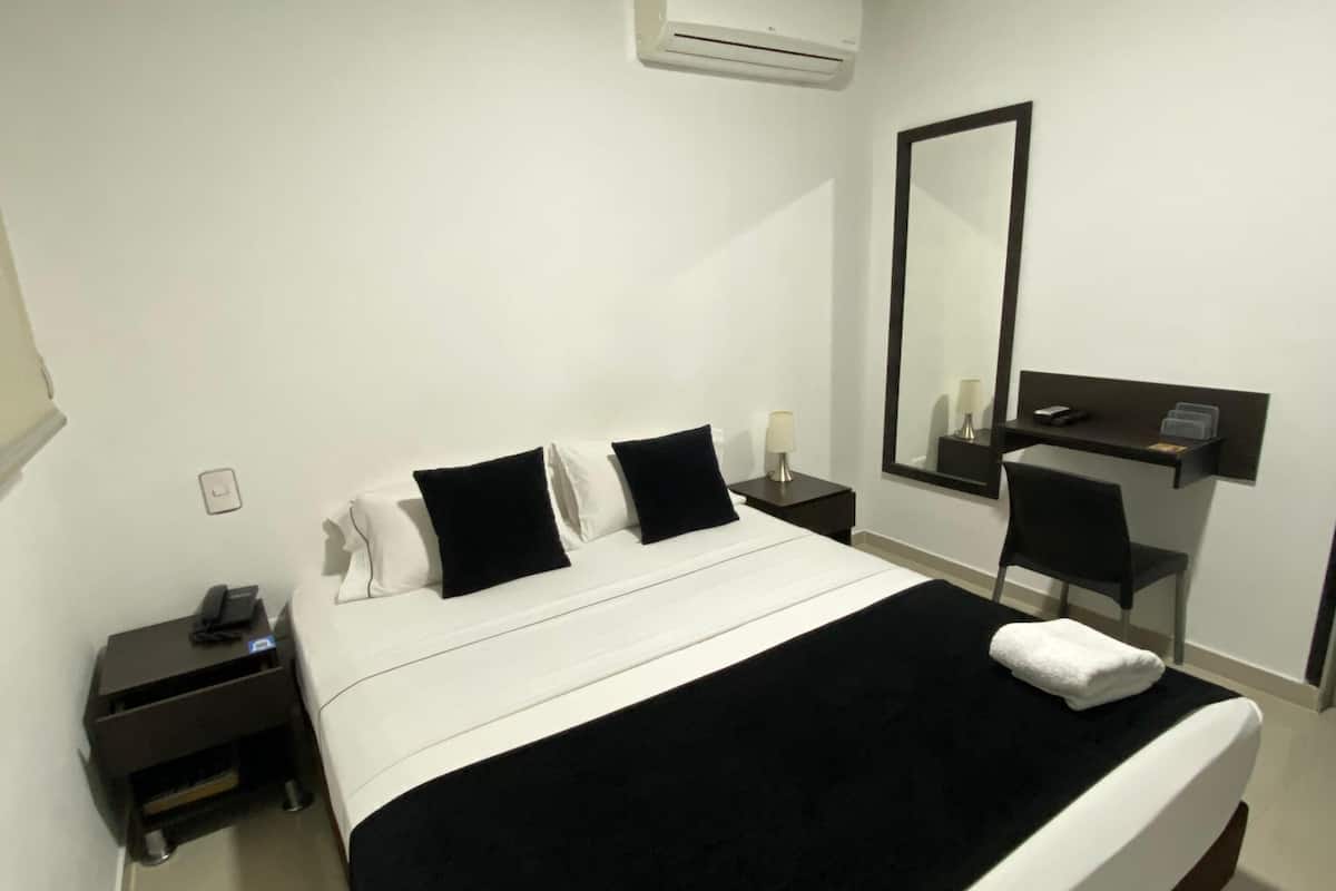 Ein Hotelzimmer mit einem großen Bett, einem Nachttisch, einem Schreibtisch, einem Stuhl, einem Spiegel und einer Klimaanlage.