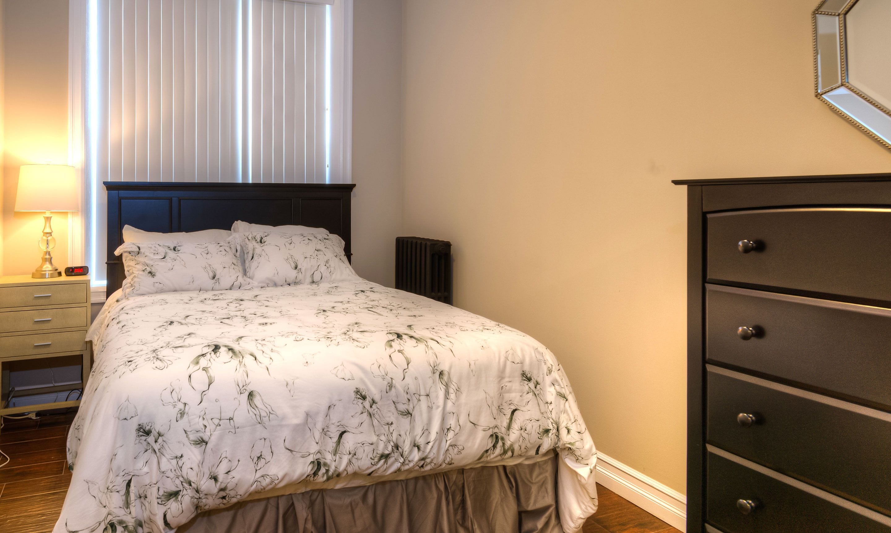Une chambre à coucher comprenant un lit, une commode, une table de chevet, une lampe et une fenêtre avec des stores.