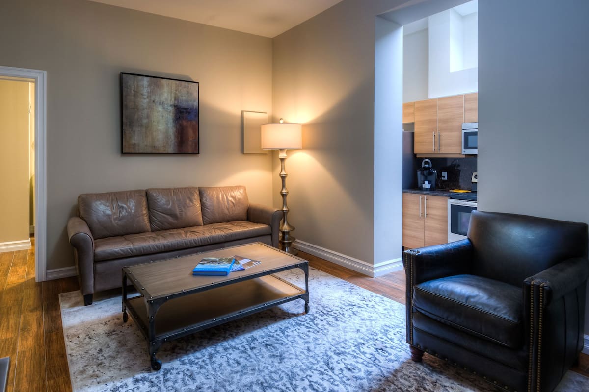 A living room with a sofa, a coffee table, a lamp, and a chair. There is a painting on the wall and a kitchen area visible through an open door.