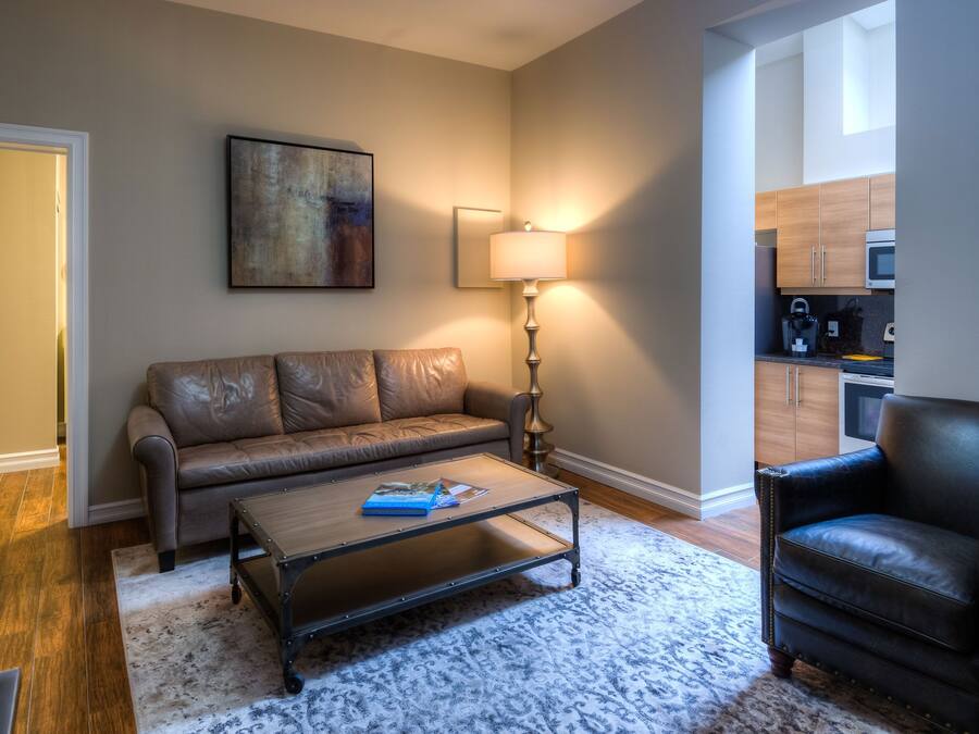 A living room with a sofa, a coffee table, a lamp, and a chair. There is a painting on the wall and a kitchen area visible through an open door.