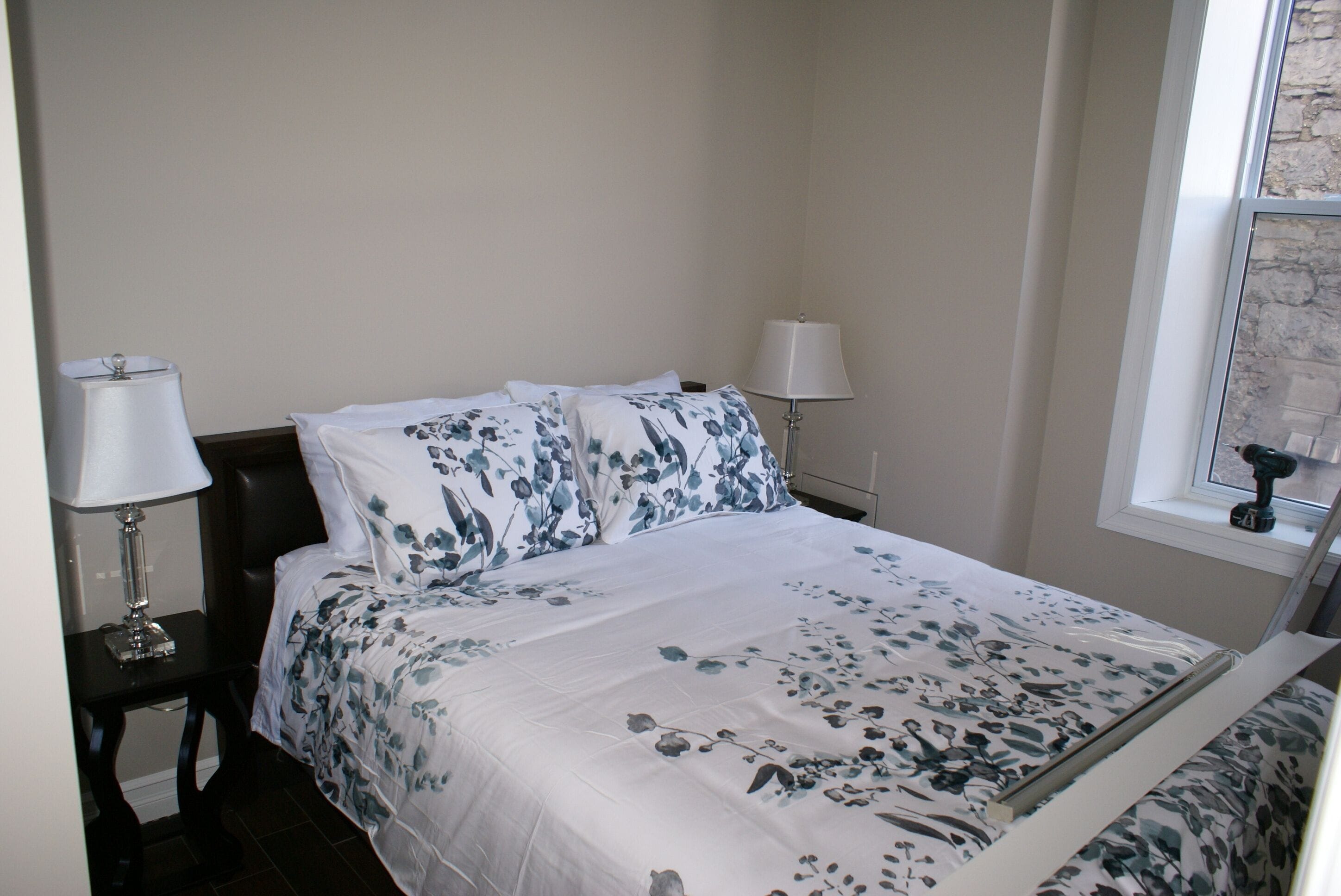 A neatly made bed with floral bedding, a bedside lamp, and a window with a view of the outside.