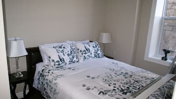 A neatly made bed with floral bedding, a bedside lamp, and a window with a view of the outside.