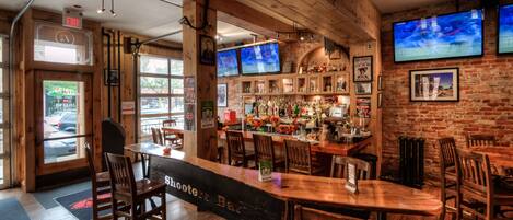 A bar area with wooden furniture, multiple screens, and a brick wall.