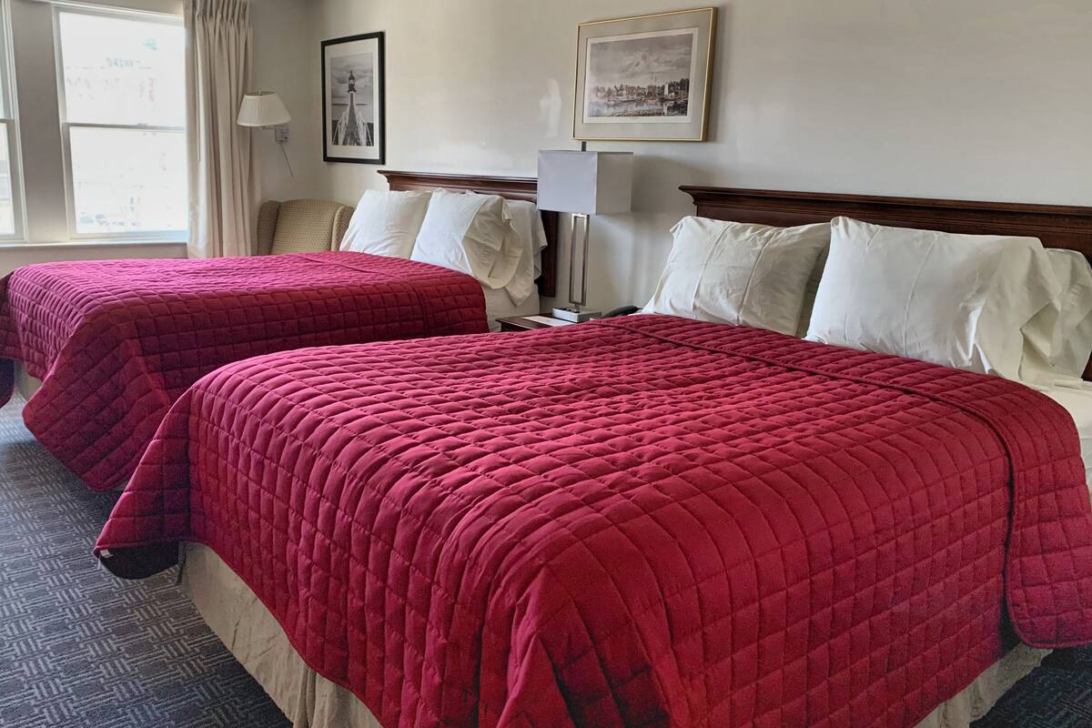 Quarto de hotel com duas camas, cada uma com colcha acolchoada vermelha, cabeceira de madeira e almofadas brancas. Há uma janela com cortinas, uma luminária e duas molduras na parede.