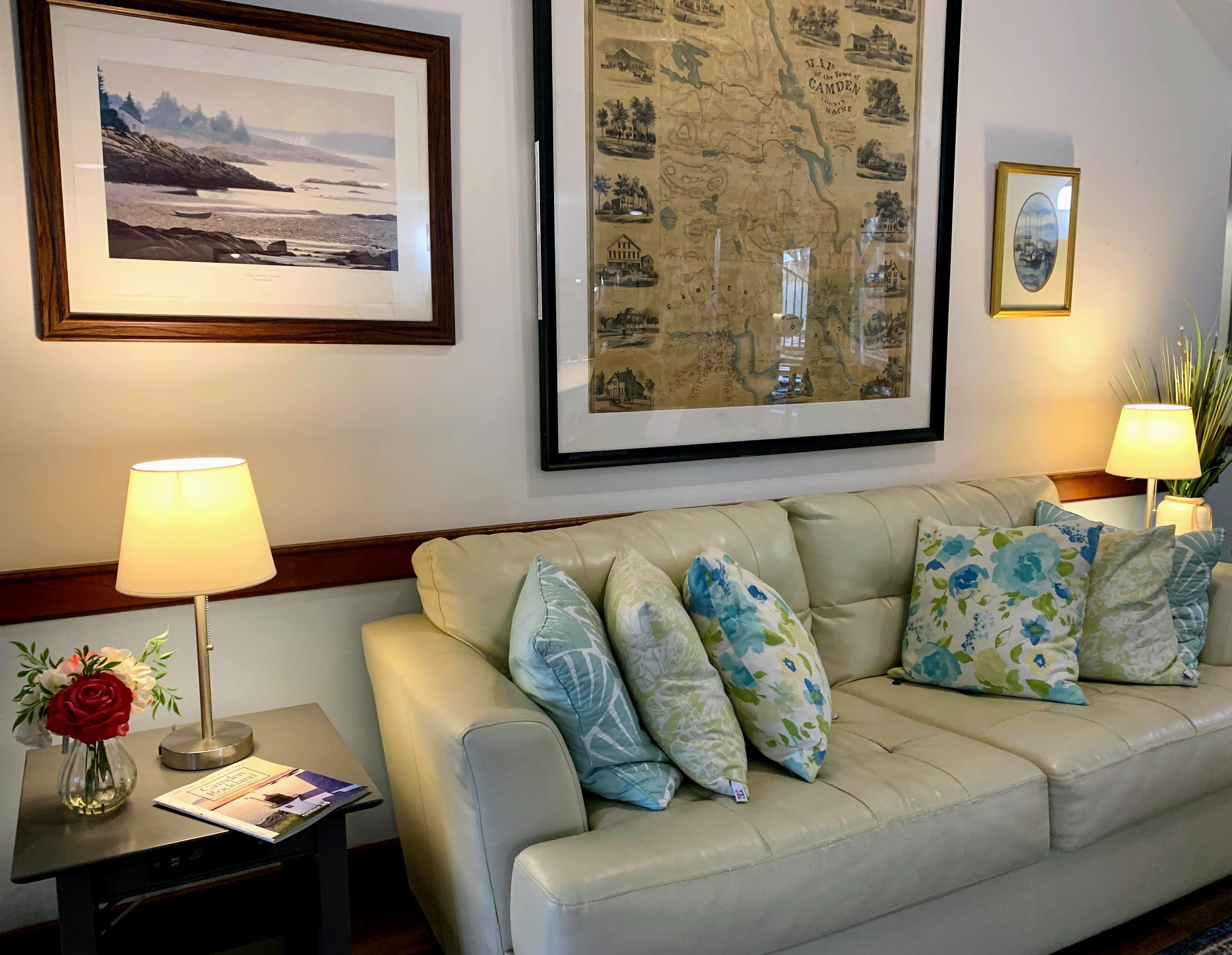 A well-lit living room with a beige sofa, floral cushions, a framed map, and a framed photograph on the wall.