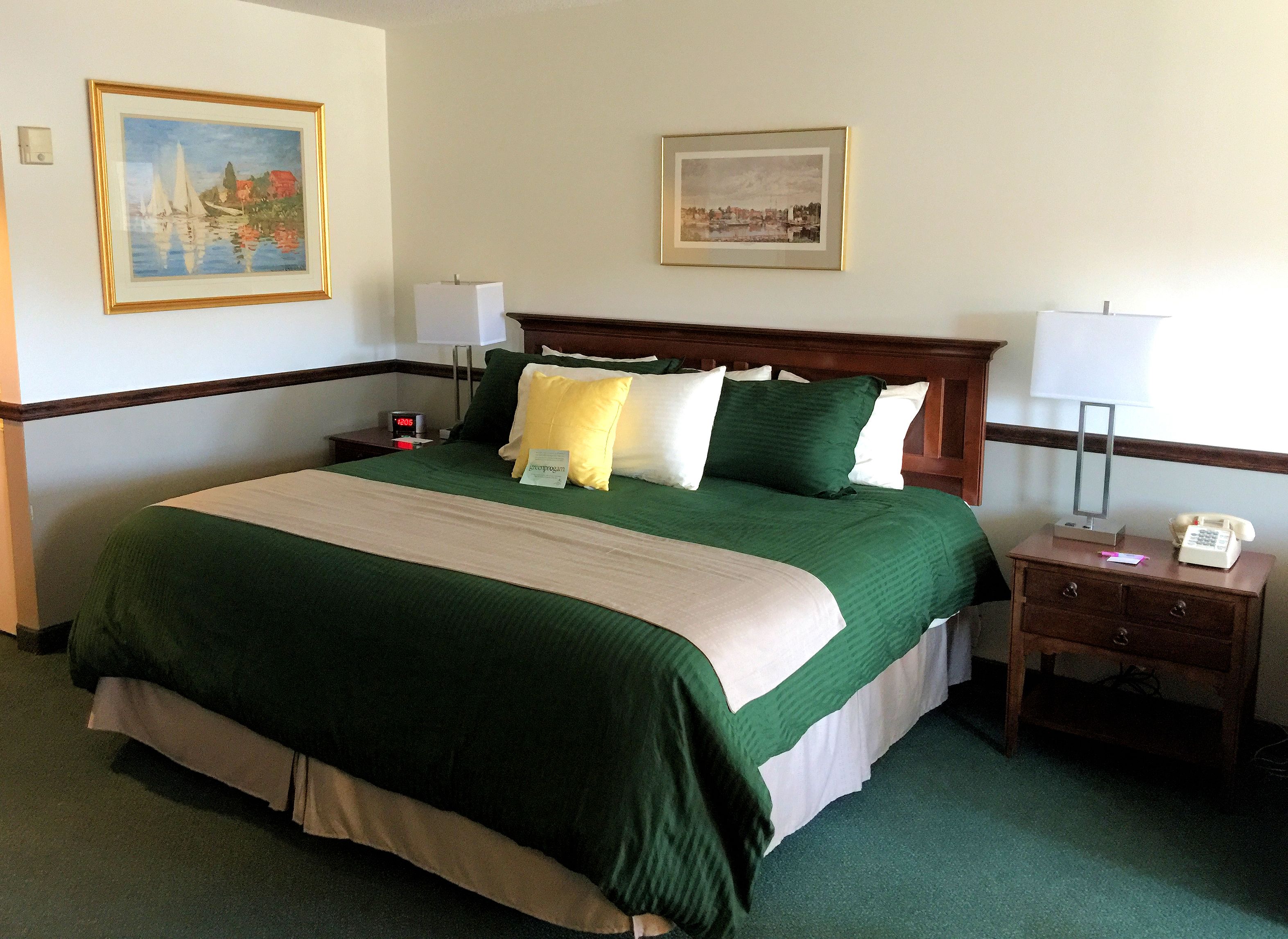 Een hotelkamer met een groot bed, beddengoed in groen en beige tinten, twee nachtkastjes met lampen, een bijzettafeltje met een klok en twee ingelijste foto's aan de muur.